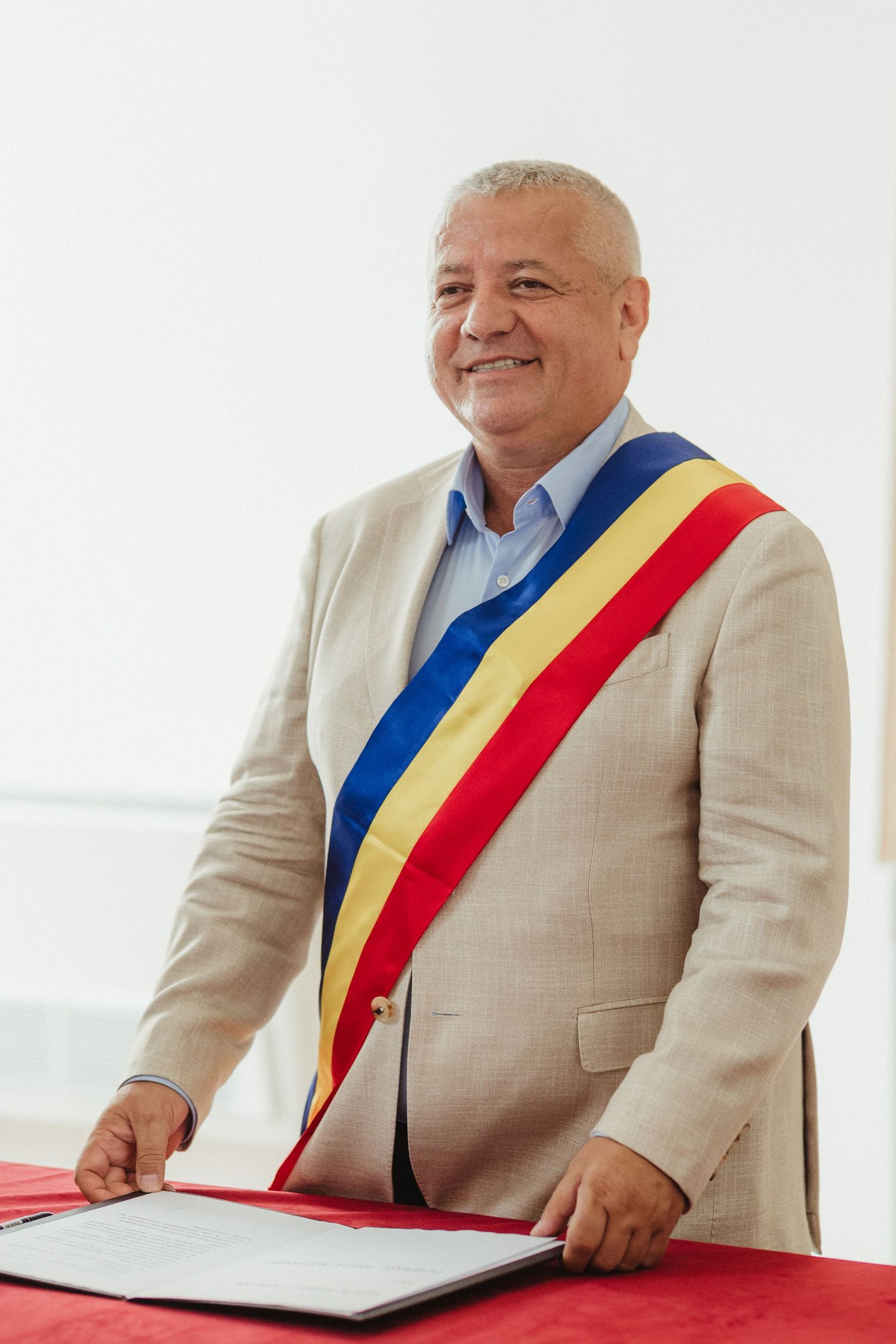 Smiling official in beige suit wearing Romanian tricolor sash standing at ceremony table.