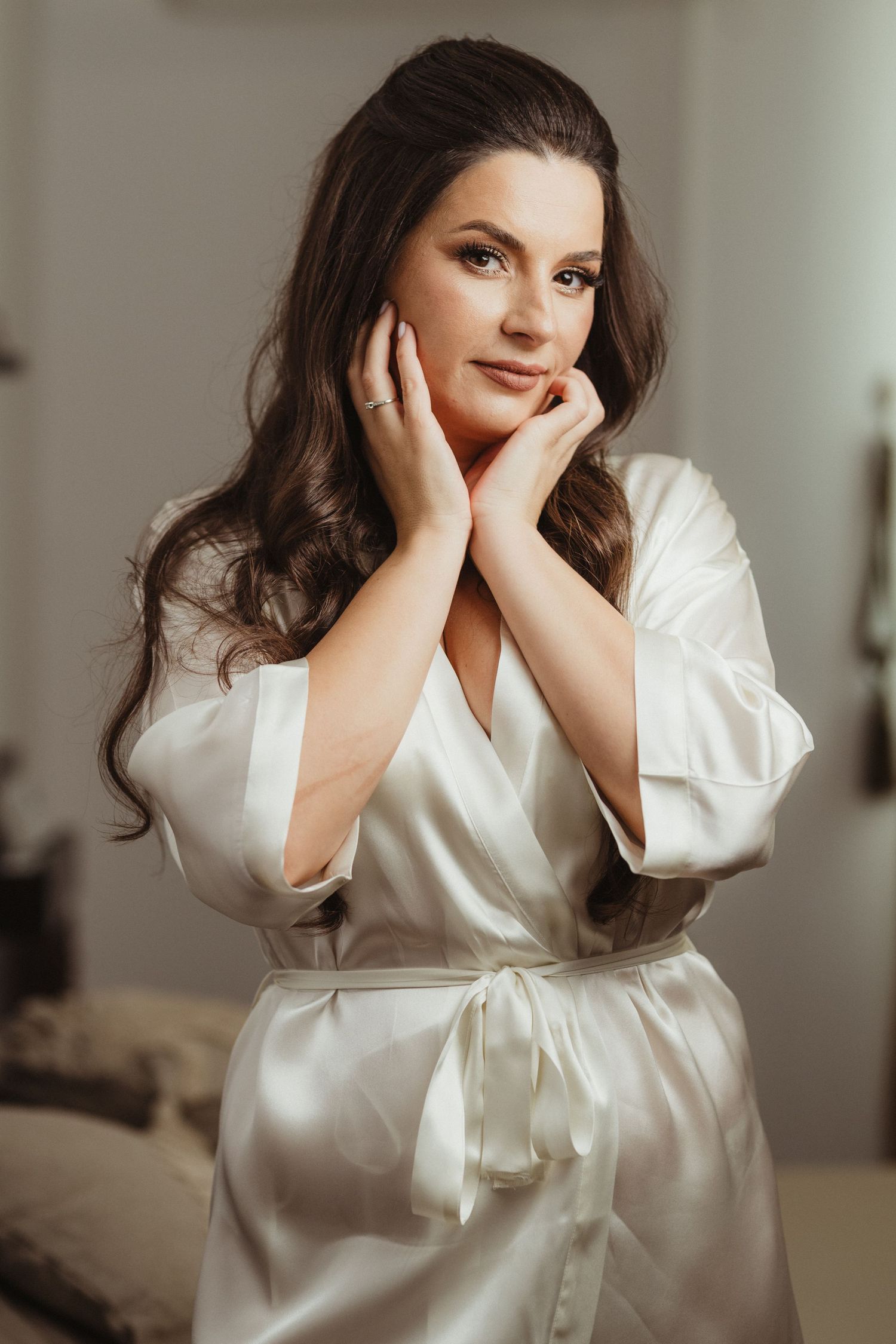 Elegant bride in white satin robe with hands near face, soft natural light, pre-wedding morning preparation.
