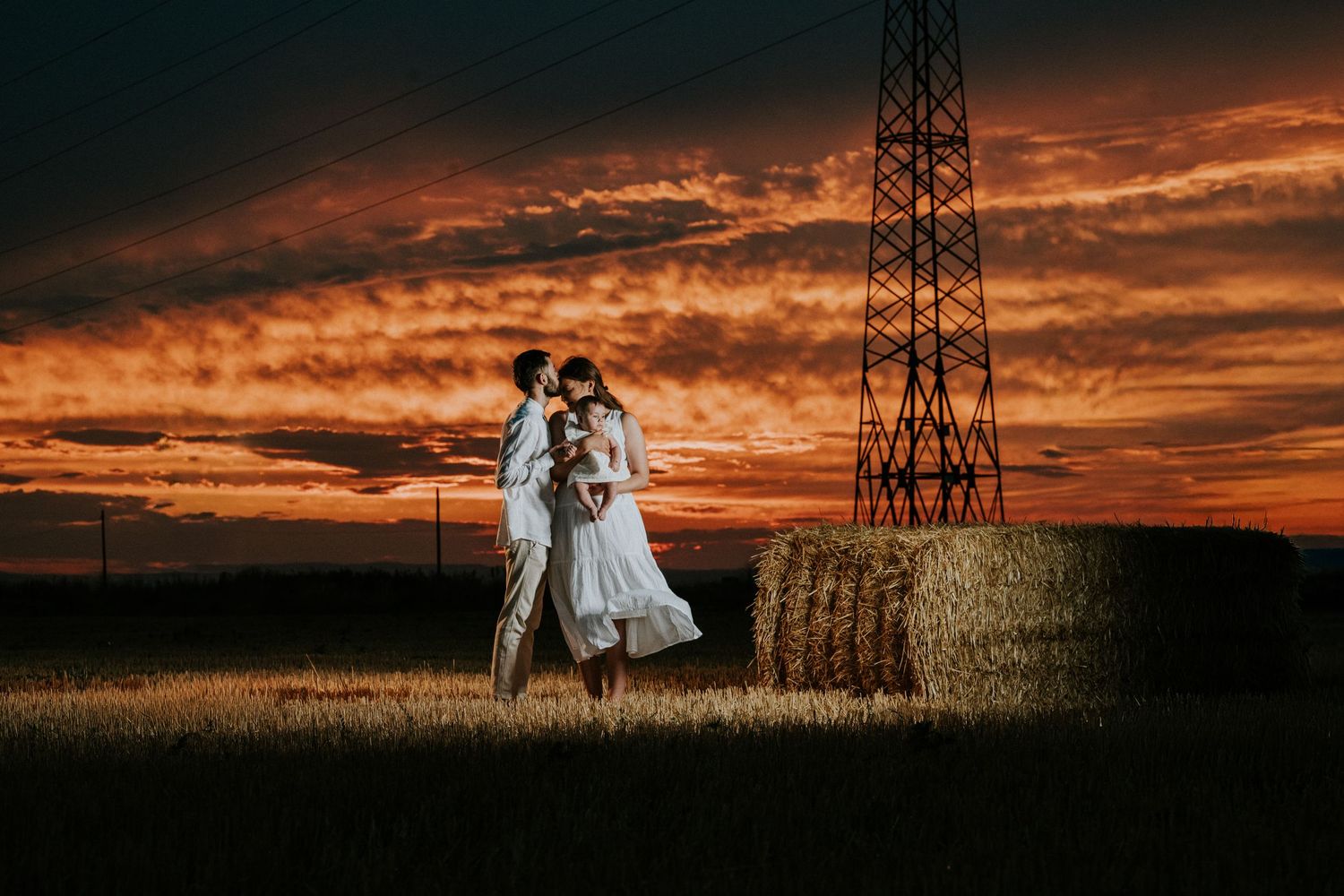 Wedding couple kissing beside hay bale in wheat field under dramatic red sunset sky.