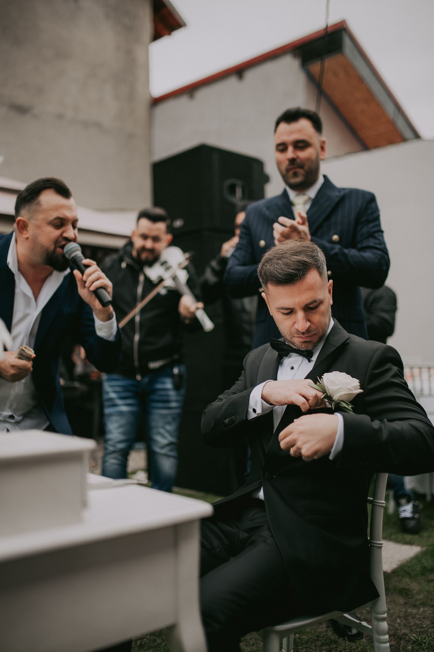 Groom in black tuxedo pinning boutonniere while musicians perform during outdoor wedding preparation.
