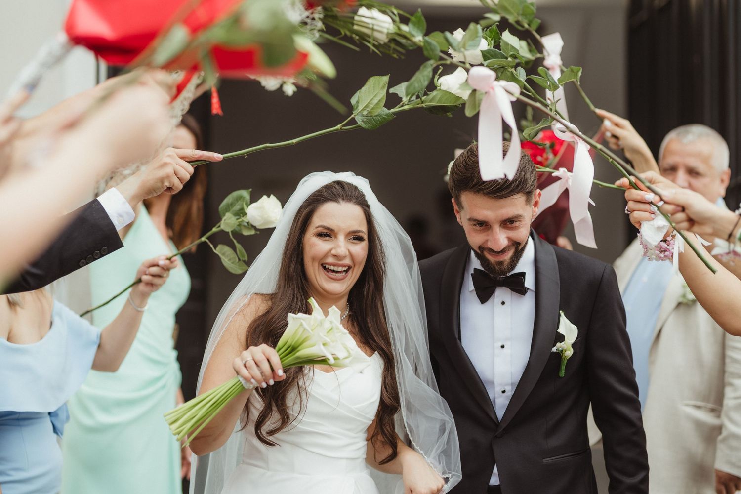 Joyful newlyweds exit church under arch of roses held by celebrating wedding guests.