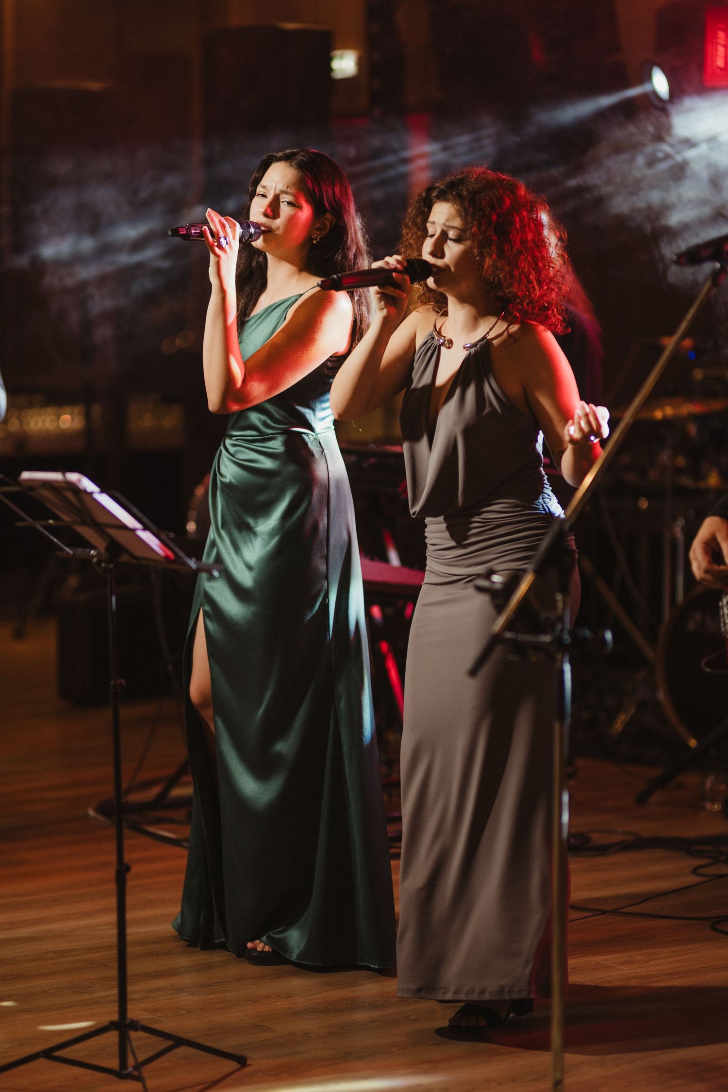 Two women singing duet on stage, one in green satin gown and one in gray dress.