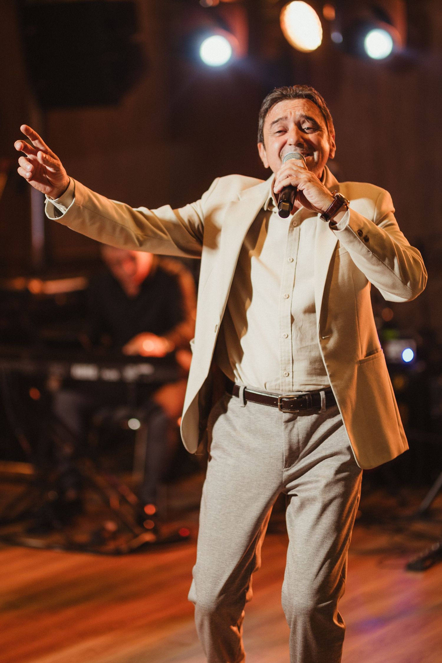 Male singer in beige blazer performing passionately on stage with arm outstretched.