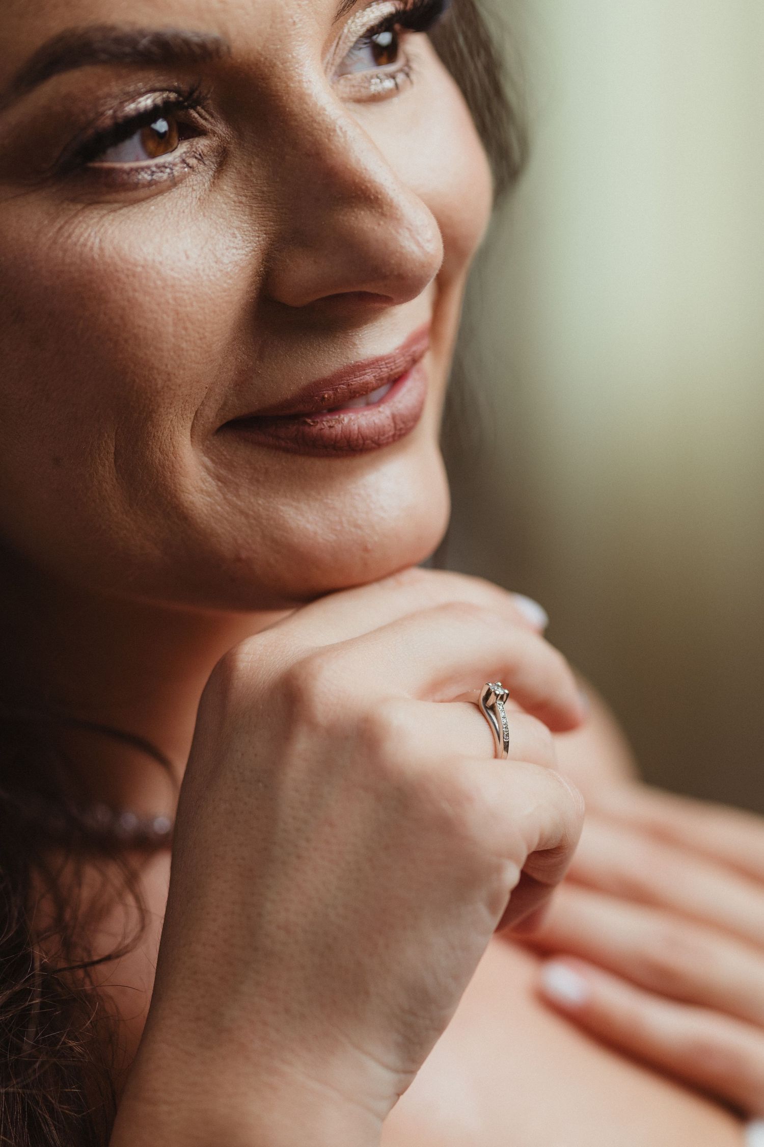 Close-up of smiling bride with diamond engagement ring, soft bokeh background, natural makeup.