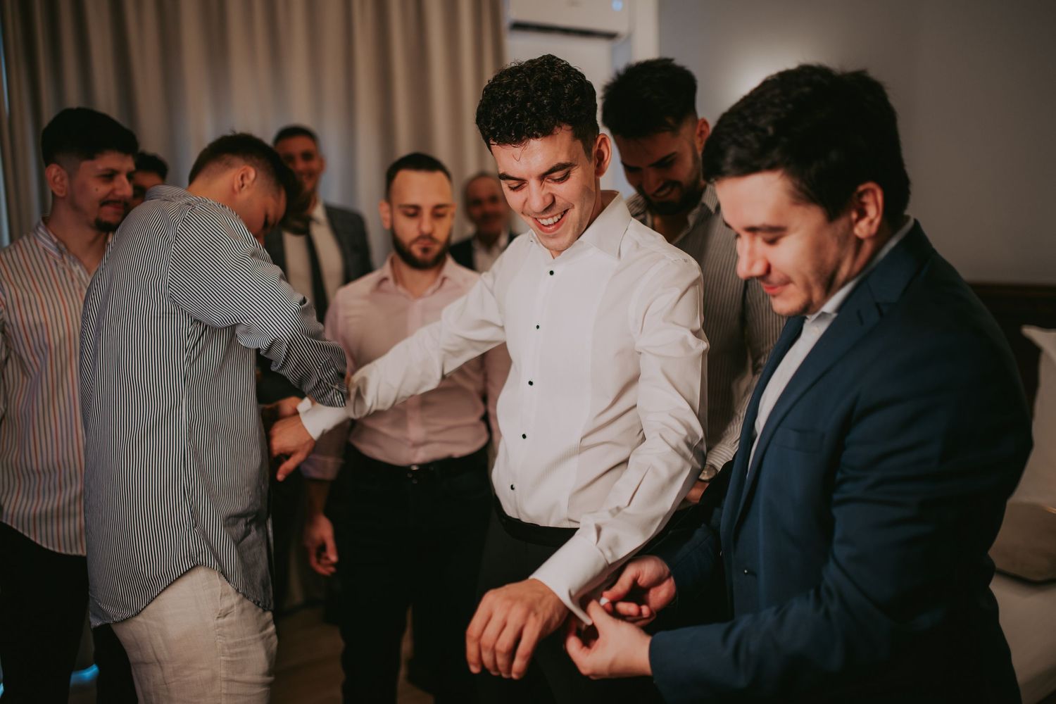 Groomsmen helping groom button his white dress shirt while laughing together indoors. Epicu Fotograf Drobeta-Turnu Severin