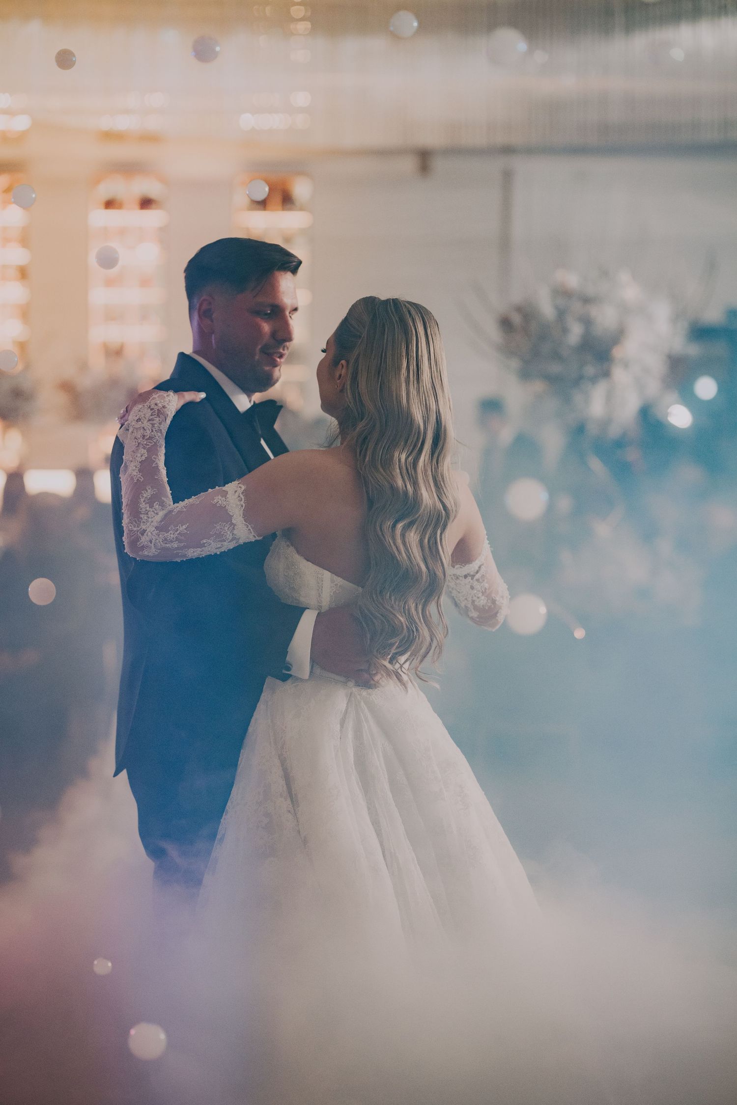 Couple sharing romantic first dance enveloped in soft fog and bokeh lights at wedding reception.
