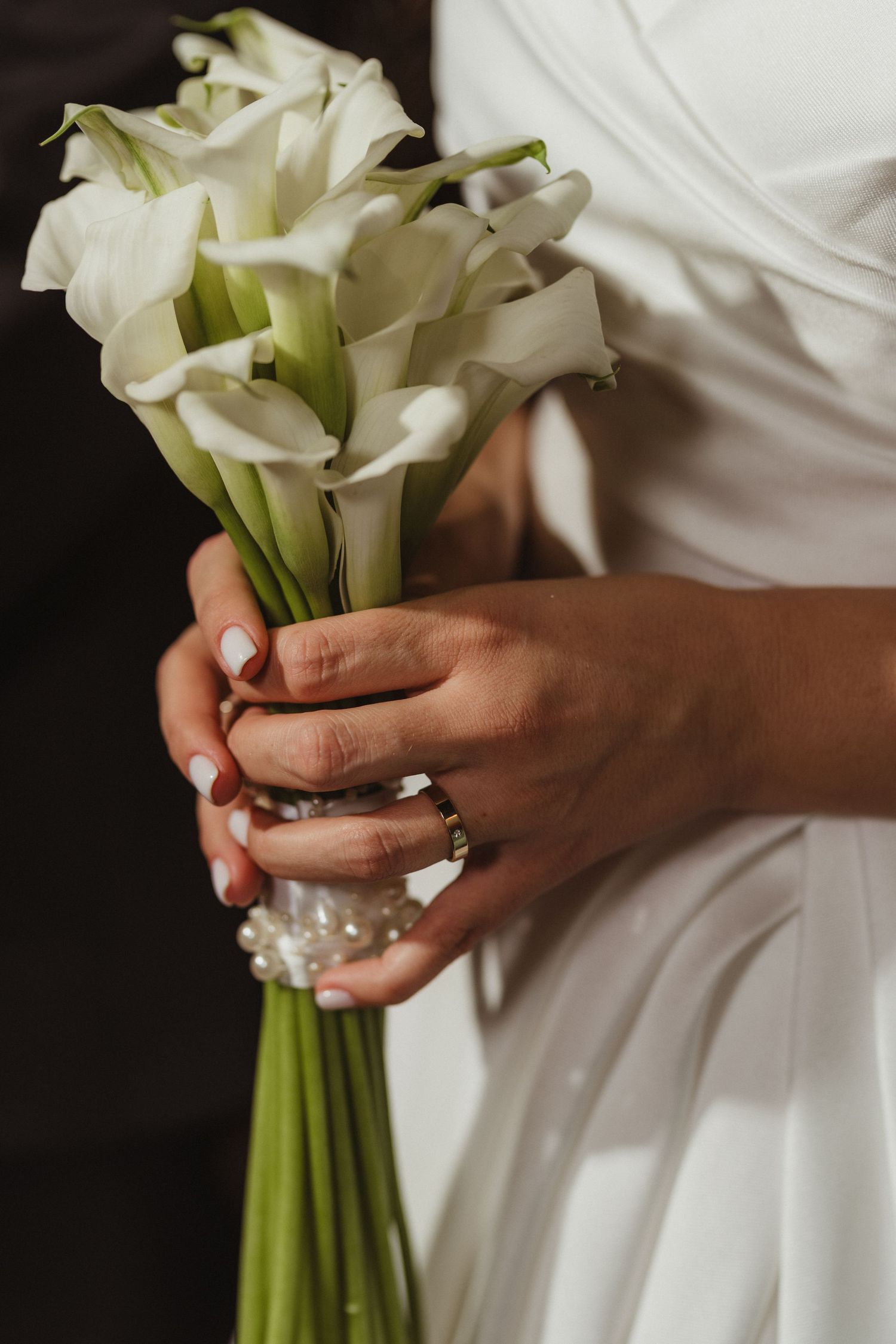 Close-up of bride holding elegant white calla lily wedding bouquet with pearl bracelet and engagement ring.