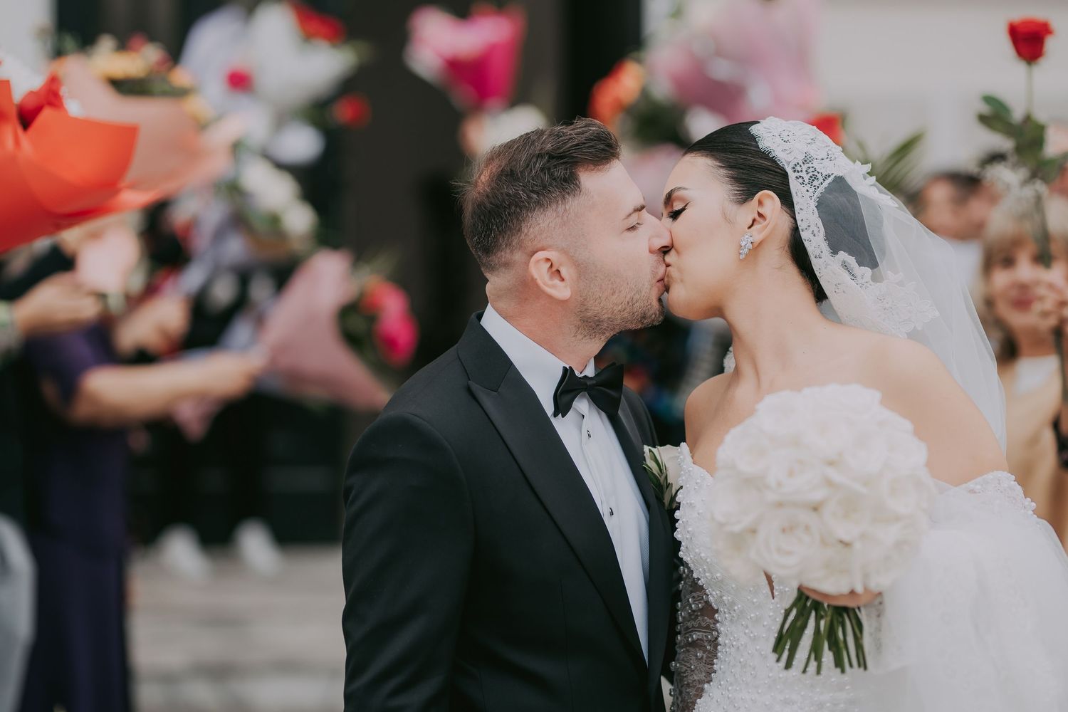 Newlyweds share first kiss outdoors surrounded by guests holding colorful flowers.