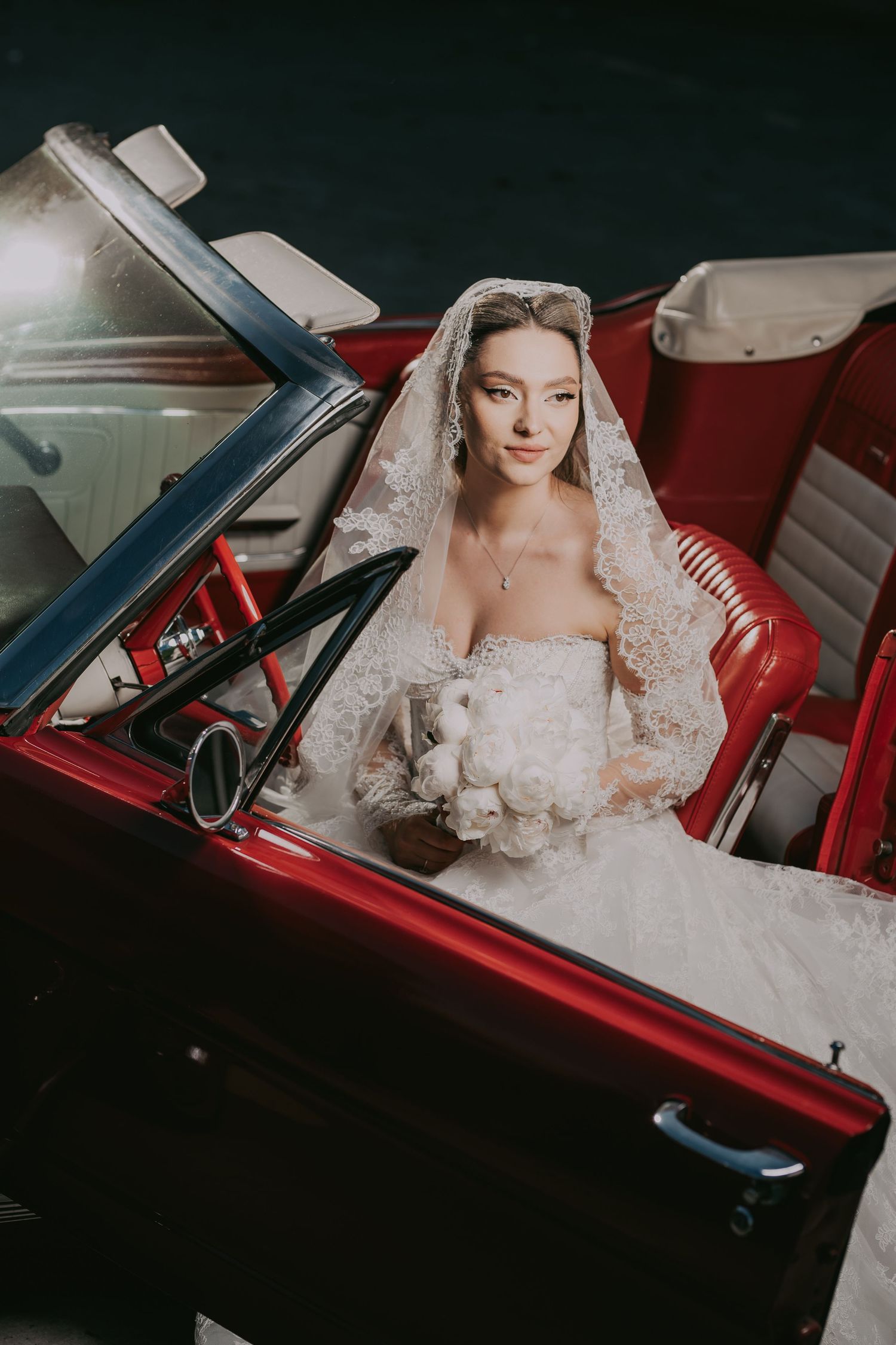 Elegant bride in lace wedding dress and veil sitting in a vintage red convertible car holding white bouquet.