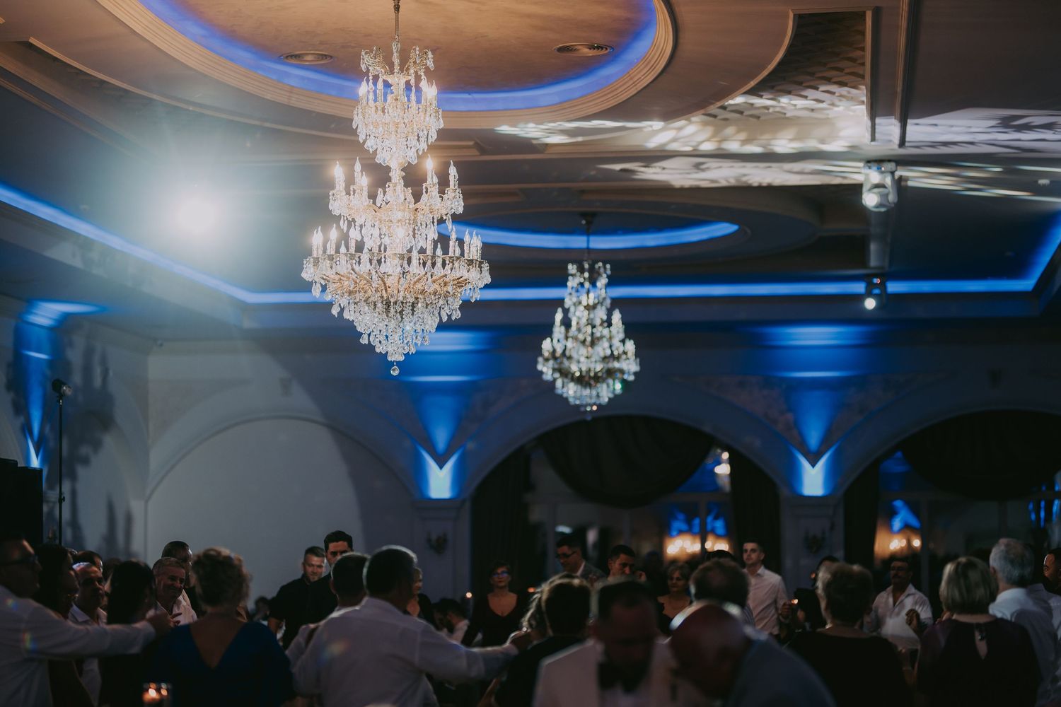 Elegant wedding reception hall with crystal chandeliers and blue uplighting as guests dance below. Epicu Fotograf Drobeta-Turnu Severin