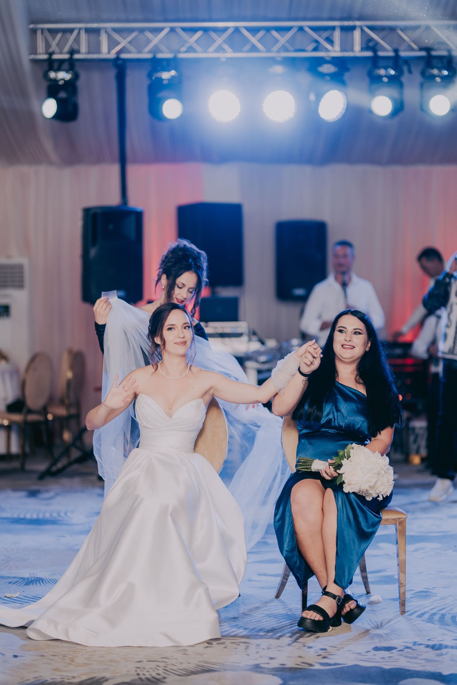 Bride in white gown and bridesmaid in blue dress sit together during a fun wedding reception game on stage. Epicu Fotograf Drobeta-Turnu Sev