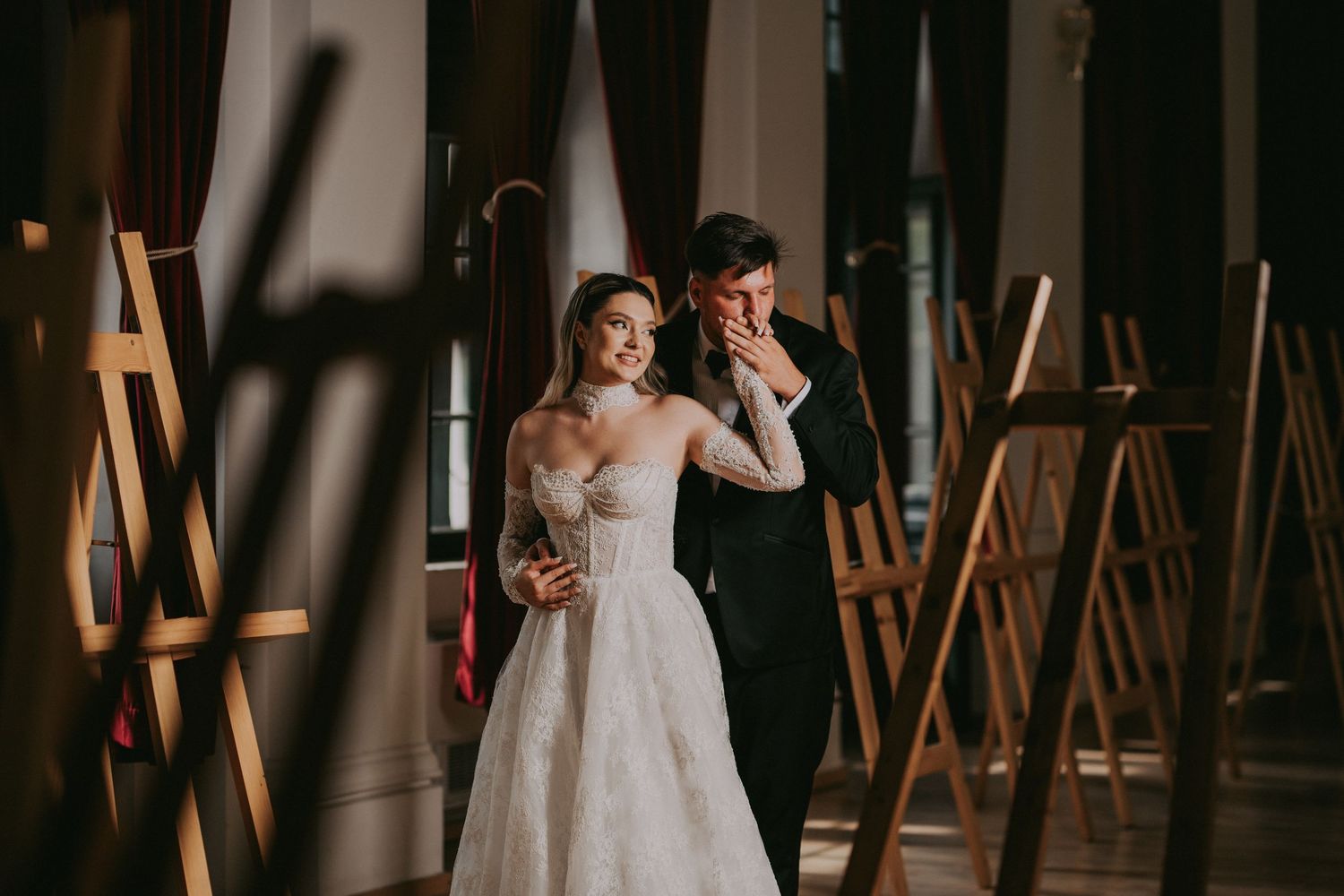 Bride and groom walk through a gallery with wooden easels in a warmly lit historic room.