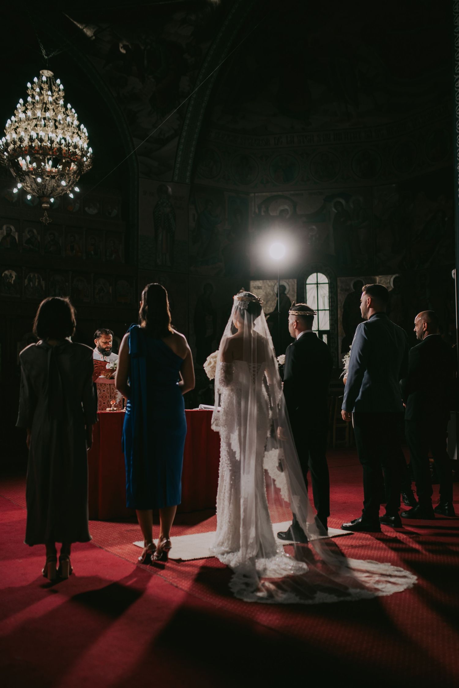 Wedding party gathered at Orthodox church altar beneath crystal chandelier with dramatic spotlight illumination.