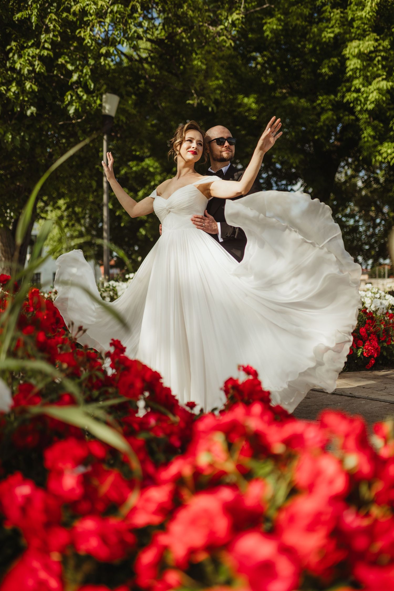 Bride in flowing white gown twirls with groom among vibrant red flowers in sunny garden setting.