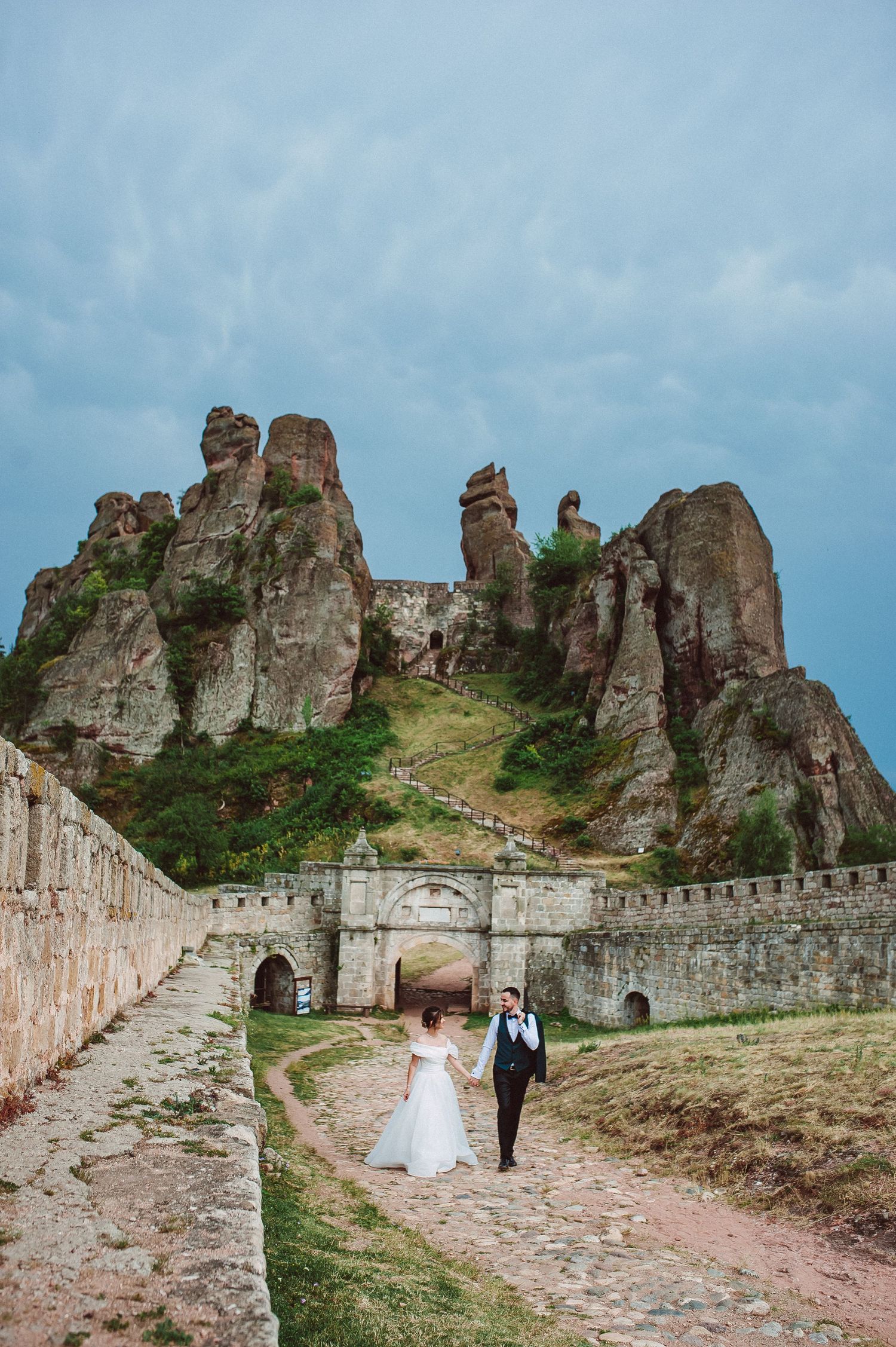 Wedding couple walking through ancient stone fortress gate with dramatic rocky cliffs and stormy sky backdrop.