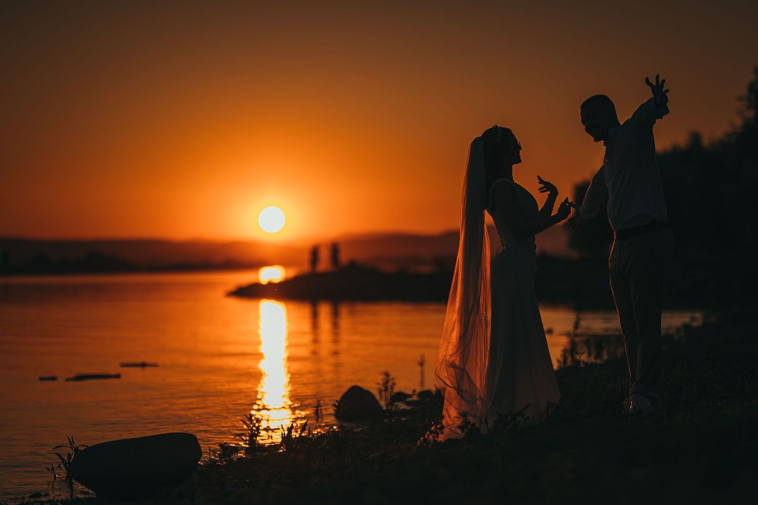 Silhouette of wedding couple holding hands by lake shore during stunning golden sunset.