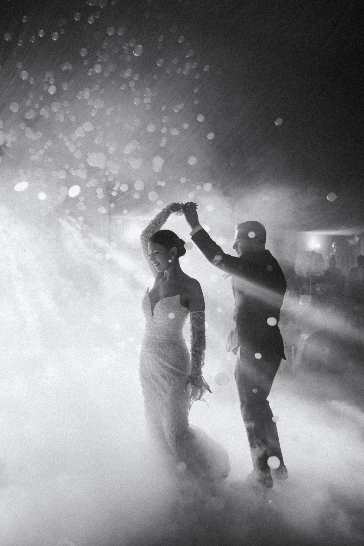 Black and white silhouette of bride and groom dancing in dramatic fog and spotlight at wedding reception.