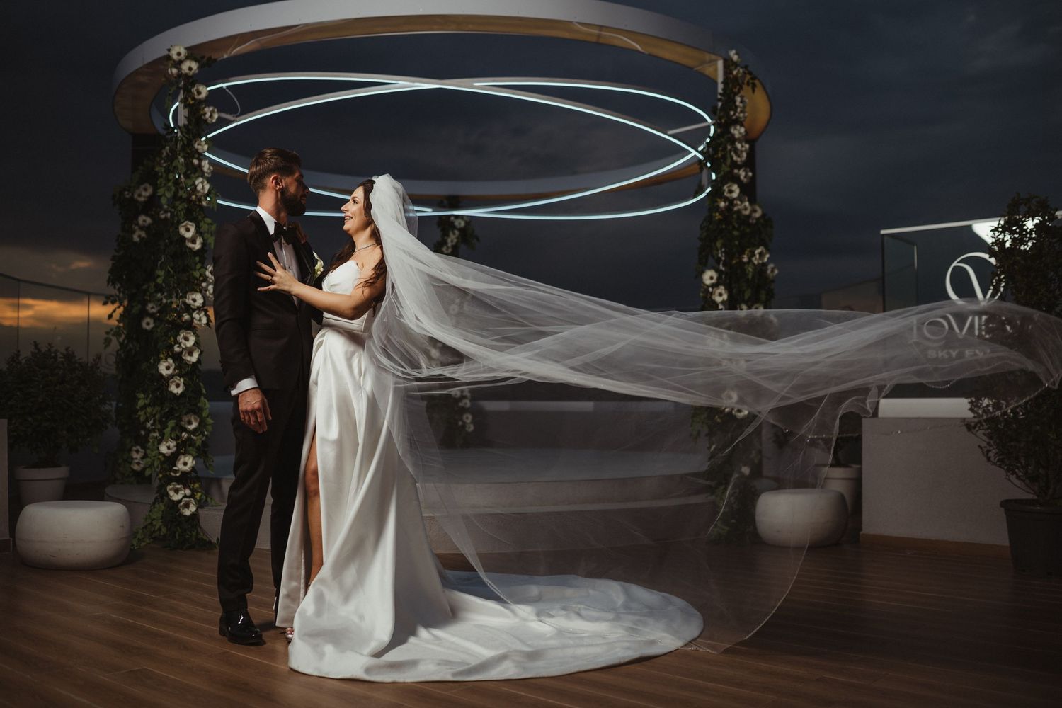 Bride and groom embrace on rooftop wedding venue at dusk, brides veil flowing dramatically in the wind under circular neon lights.