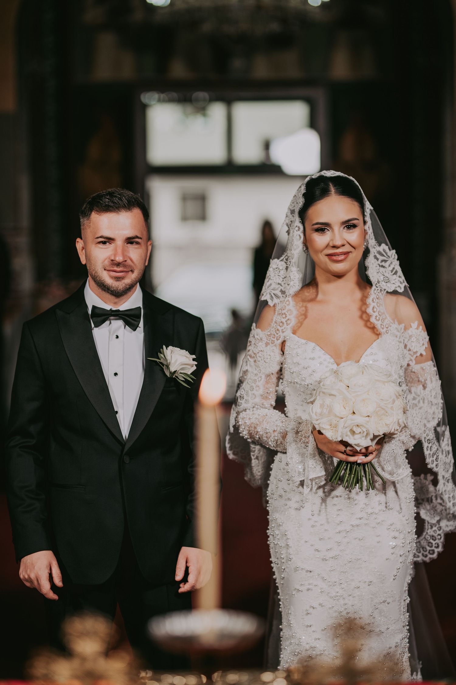 Bride in lace gown with veil and groom in black tuxedo standing together during Orthodox church wedding ceremony.