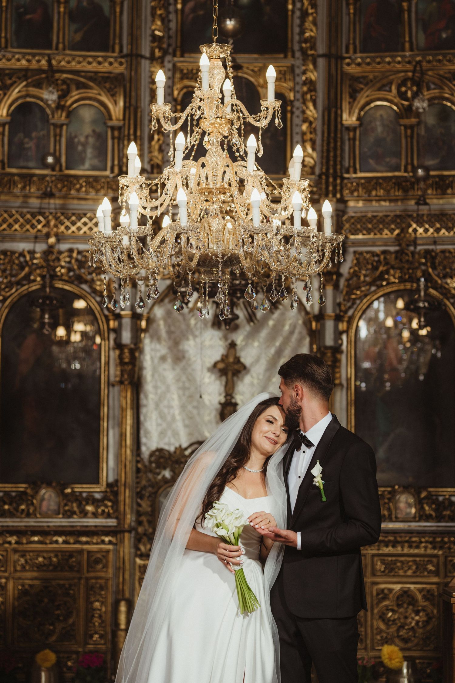 Bride and groom pose under ornate crystal chandelier in elegant golden church interior during wedding ceremony.