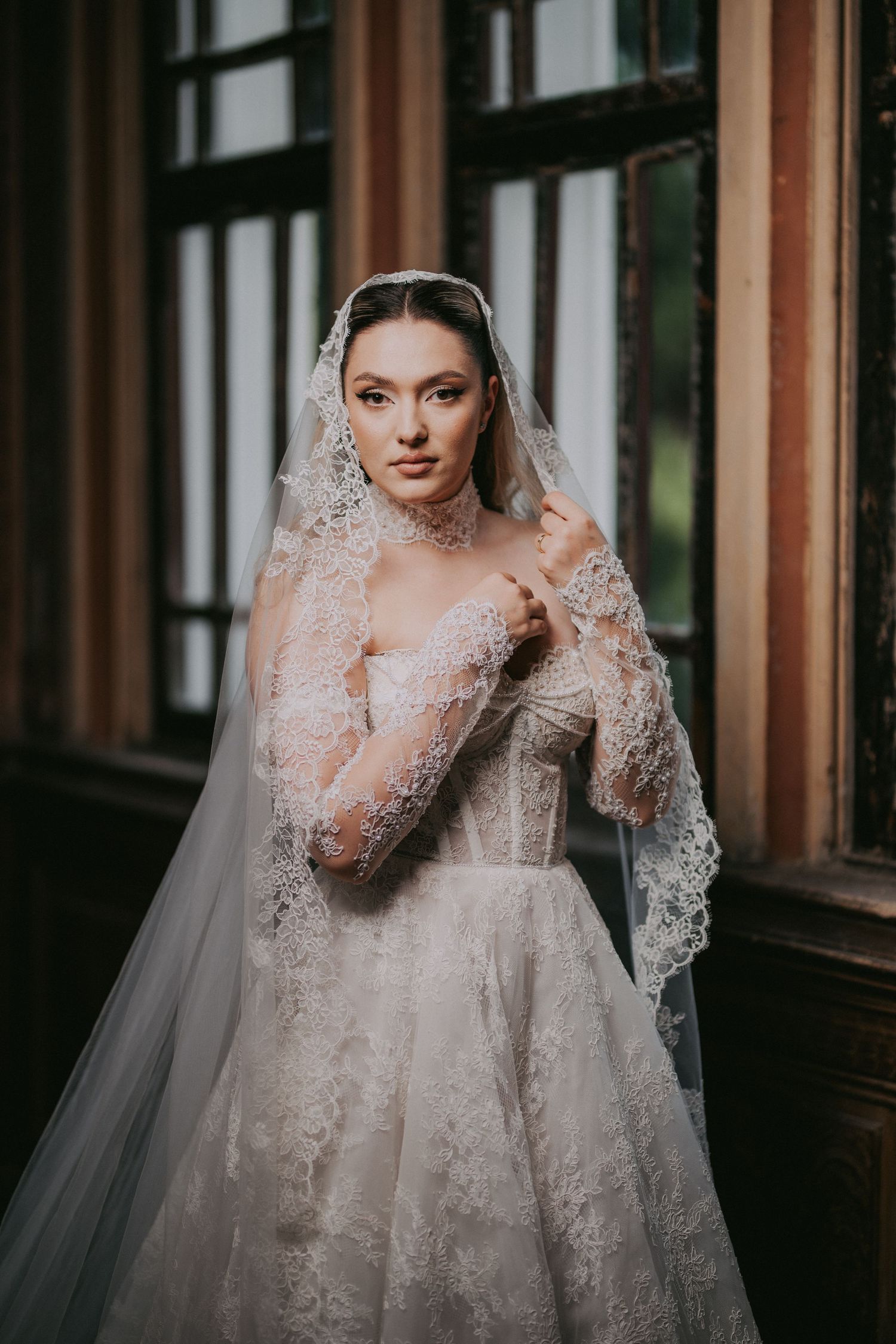 Elegant bride in lace wedding gown and cathedral veil posing gracefully by tall arched windows.