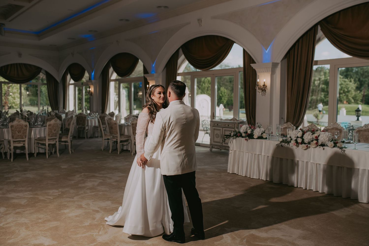 Newlyweds sharing their first dance in an elegant ballroom with arched windows and floral decorations. Epicu Fotograf Drobeta-Turnu Severin