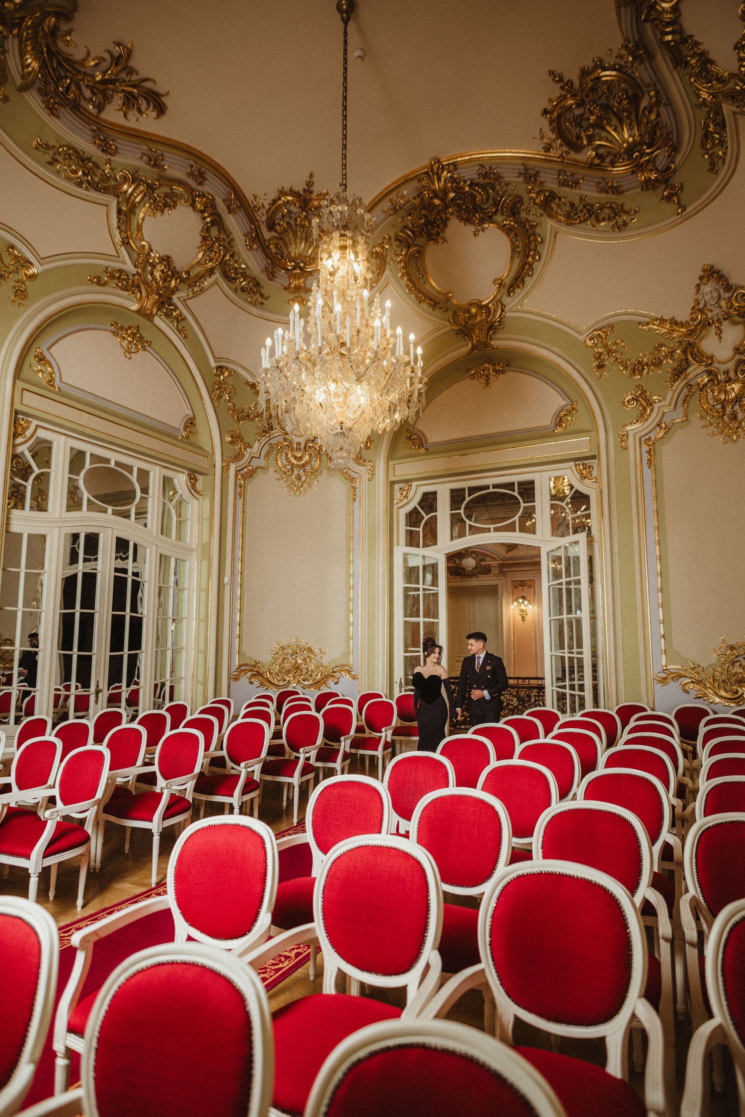 Couple standing in ornate baroque ballroom with red velvet chairs and gold ceiling decorations.