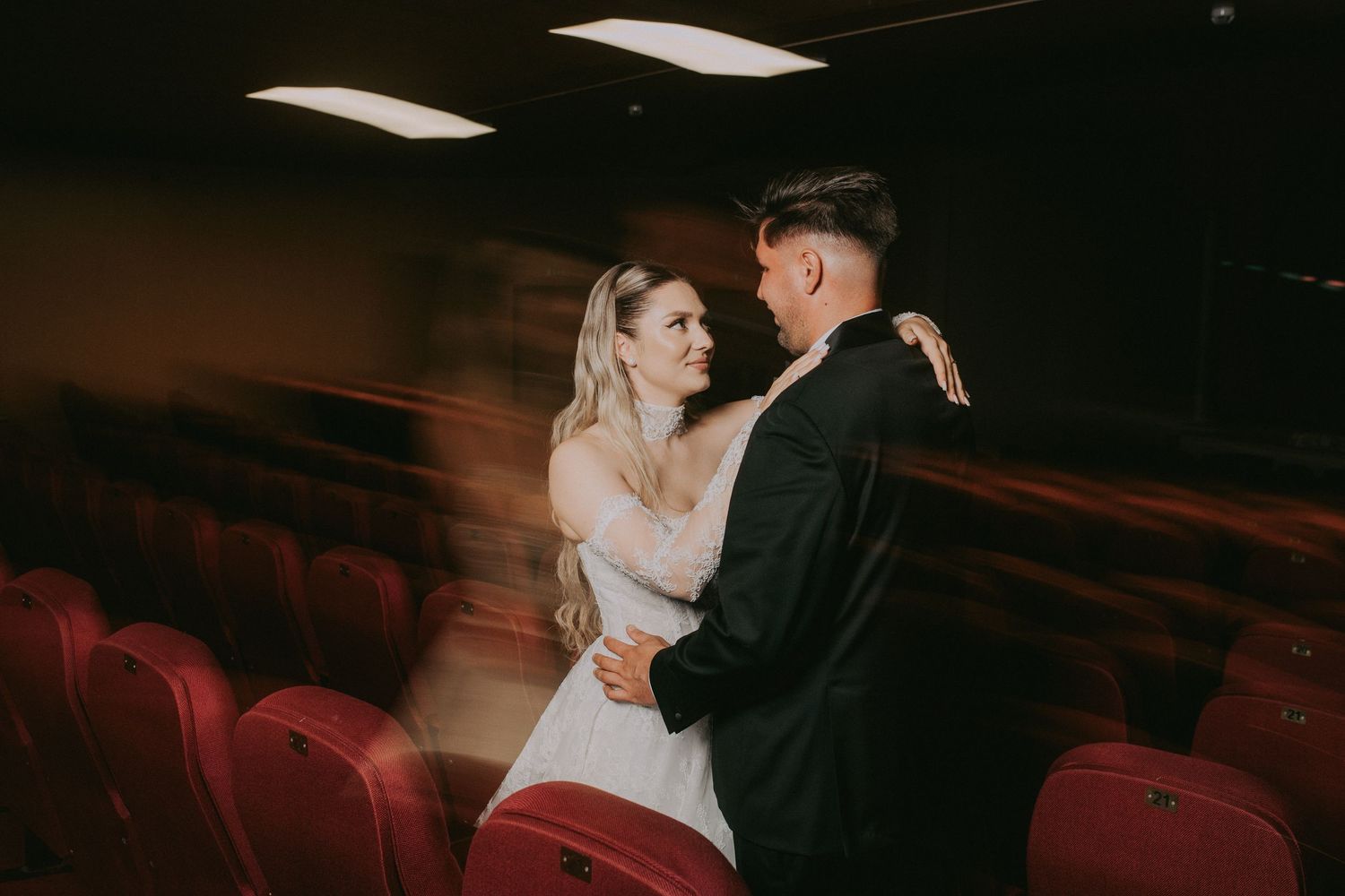 Bride and groom share a romantic dance embrace in a theater with red velvet seats.