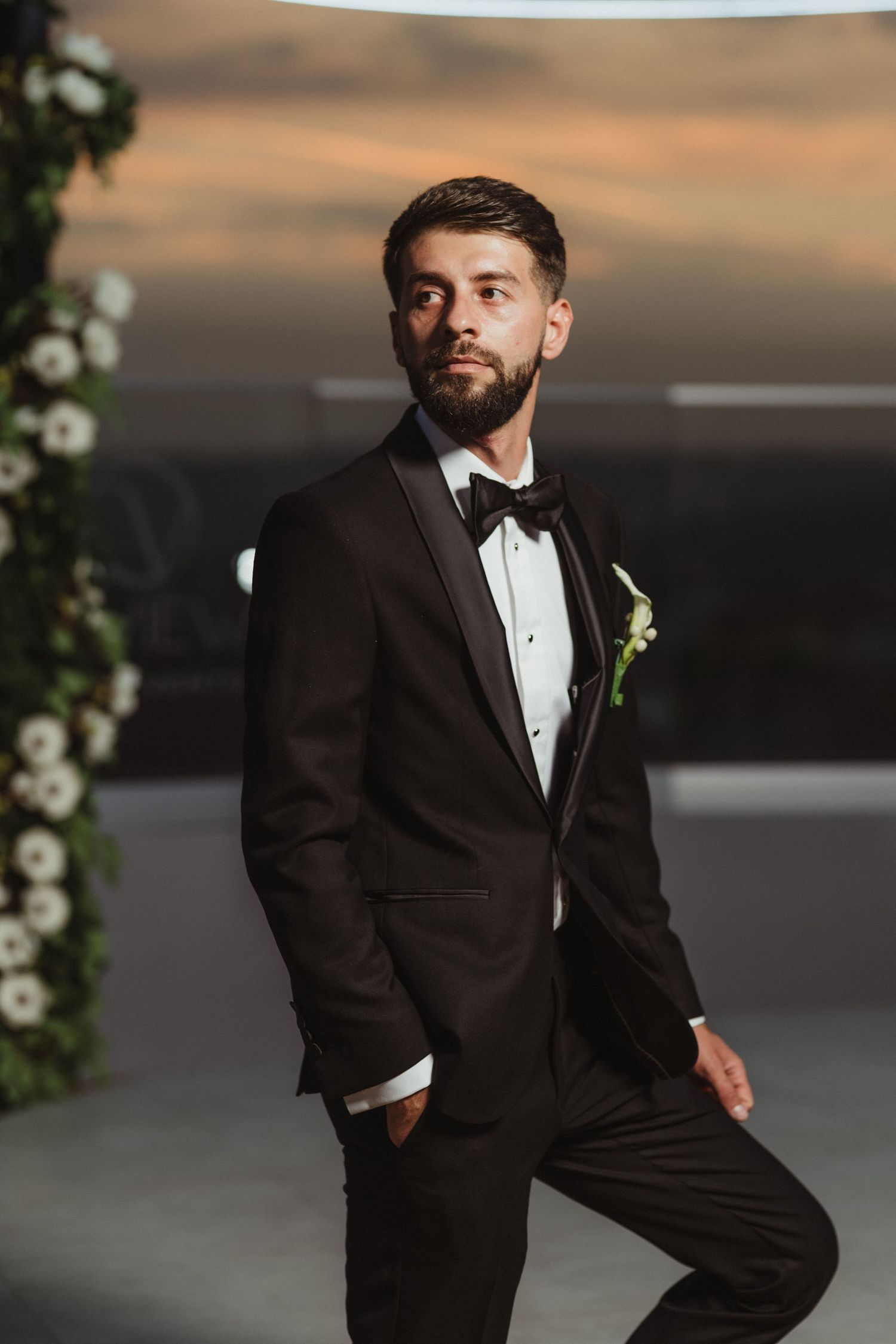 Handsome bearded groom in black tuxedo with boutonniere poses confidently at outdoor wedding venue at sunset.