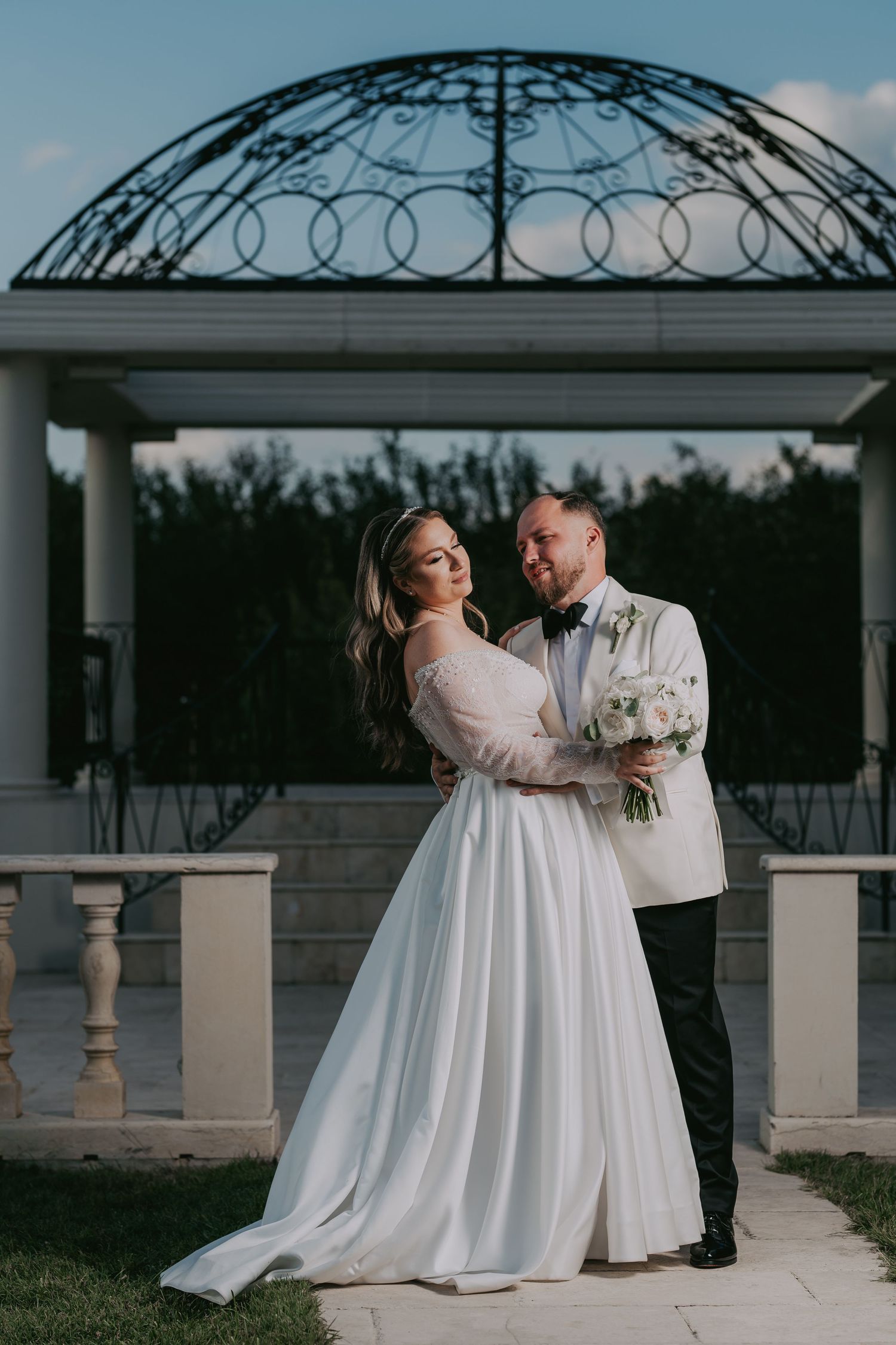 Bride and groom embrace romantically in front of decorative iron gazebo at golden hour. Epicu Fotograf Drobeta-Turnu Severin