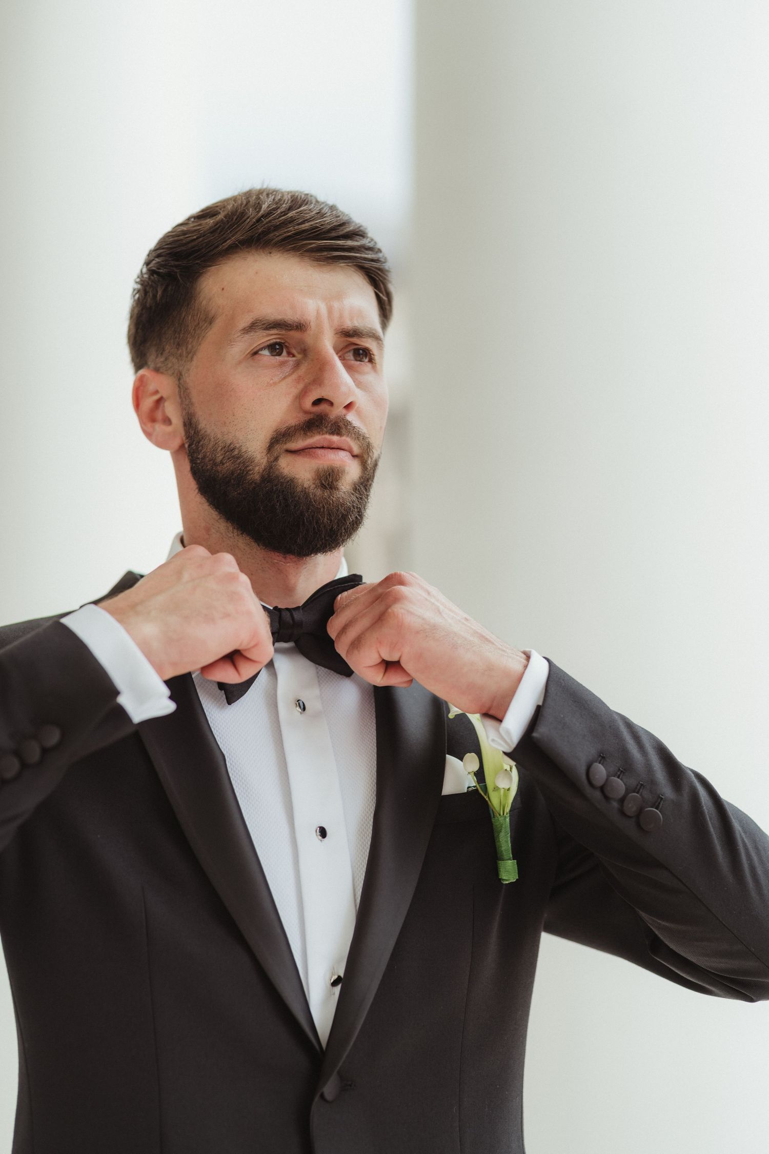 Dapper bearded groom adjusting his black bow tie while wearing a classic black tuxedo with boutonniere.