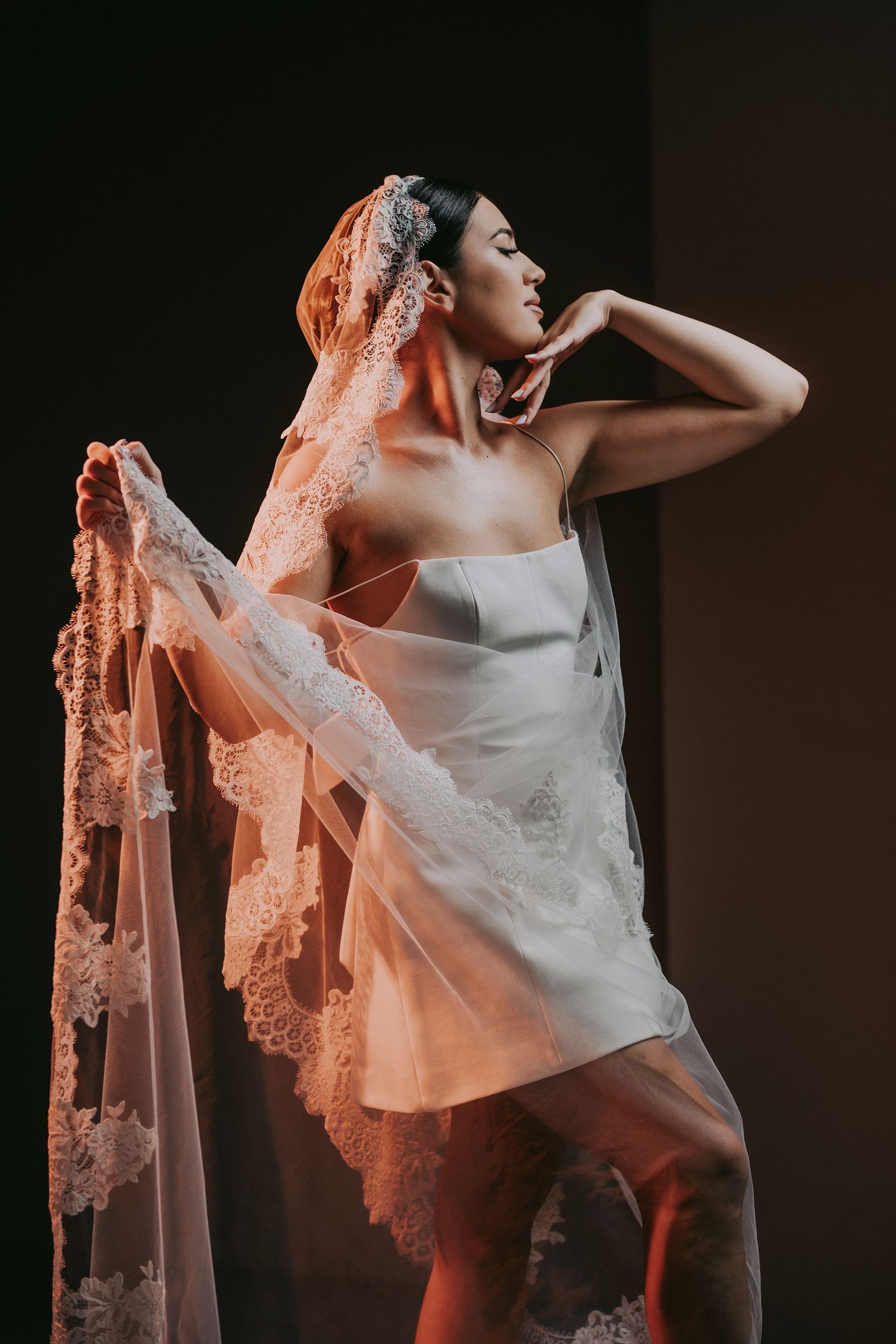 Elegant bride in short white dress posing gracefully with lace veil in dramatic warm studio lighting.