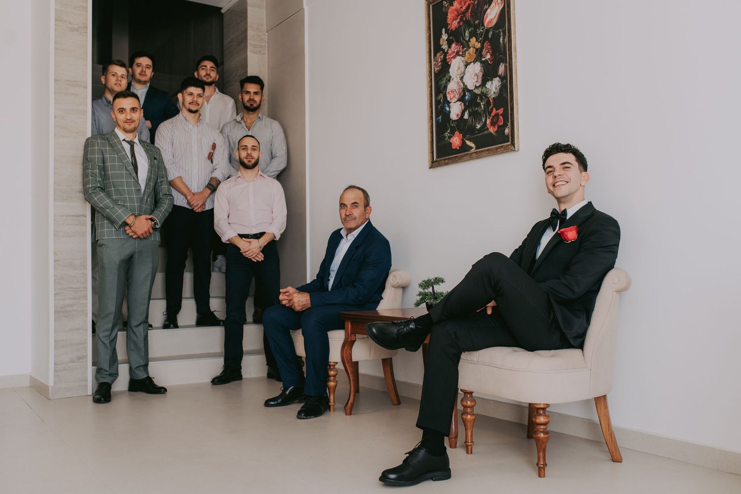 Groom in black tuxedo seated in chair surrounded by groomsmen in formal attire. Epicu Fotograf Drobeta-Turnu Severin