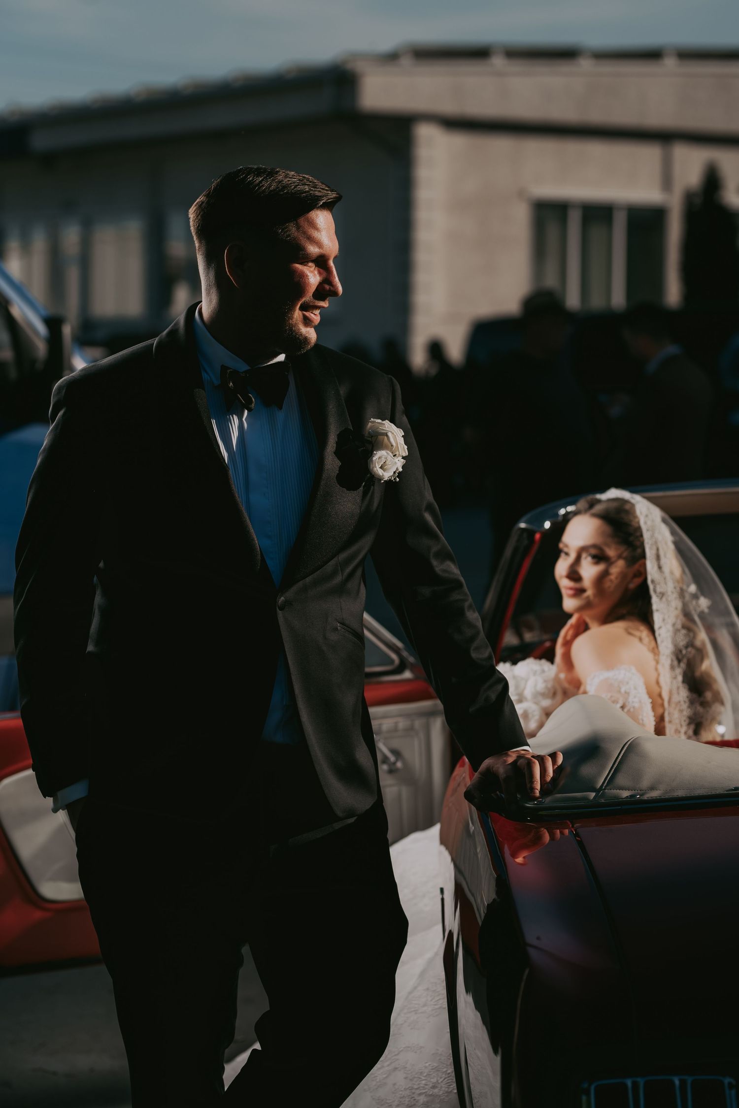 Stylish groom smiling beside red classic car with bride inside.