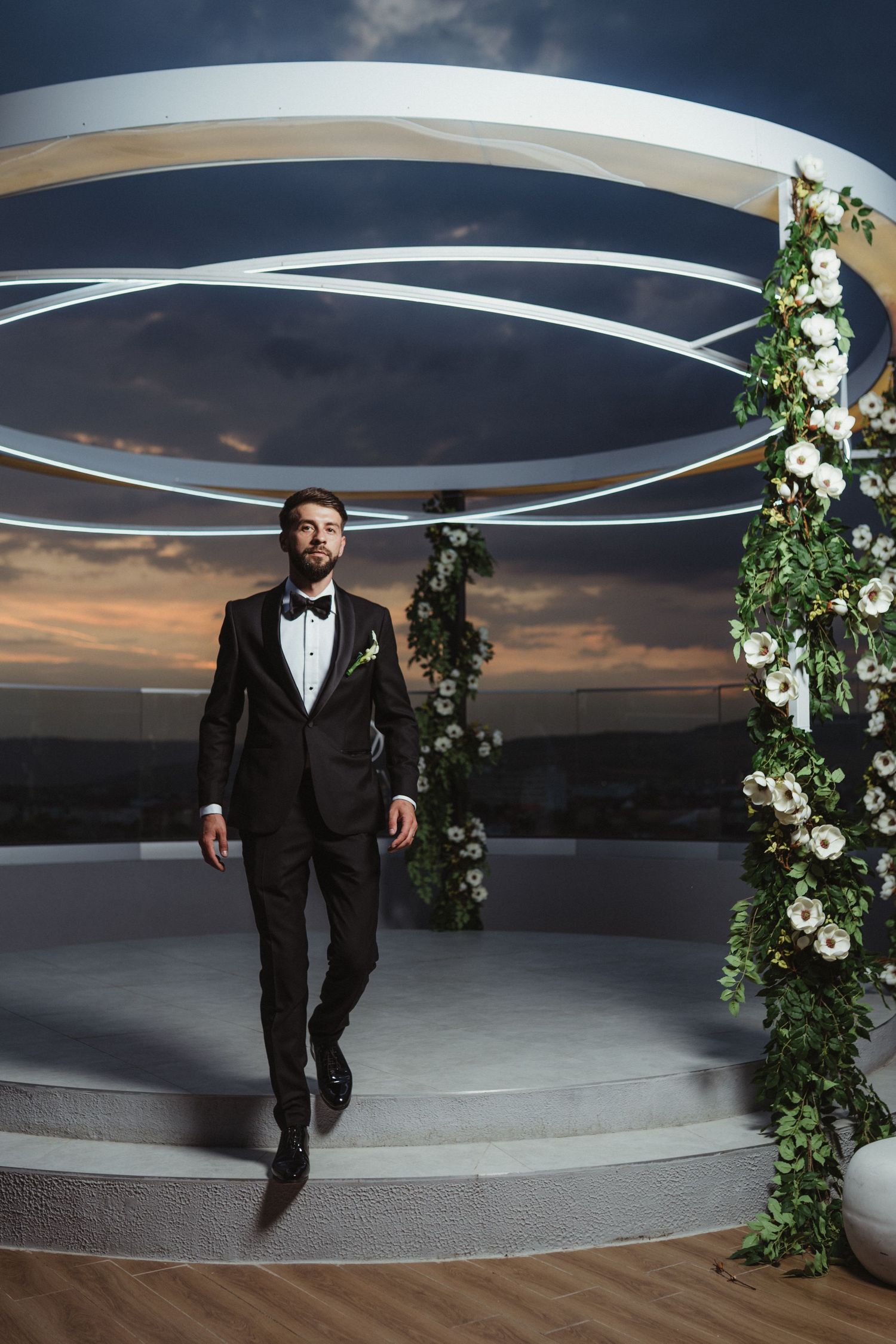 Elegant groom in black tuxedo poses at rooftop wedding ceremony space adorned with white floral arch at sunset.