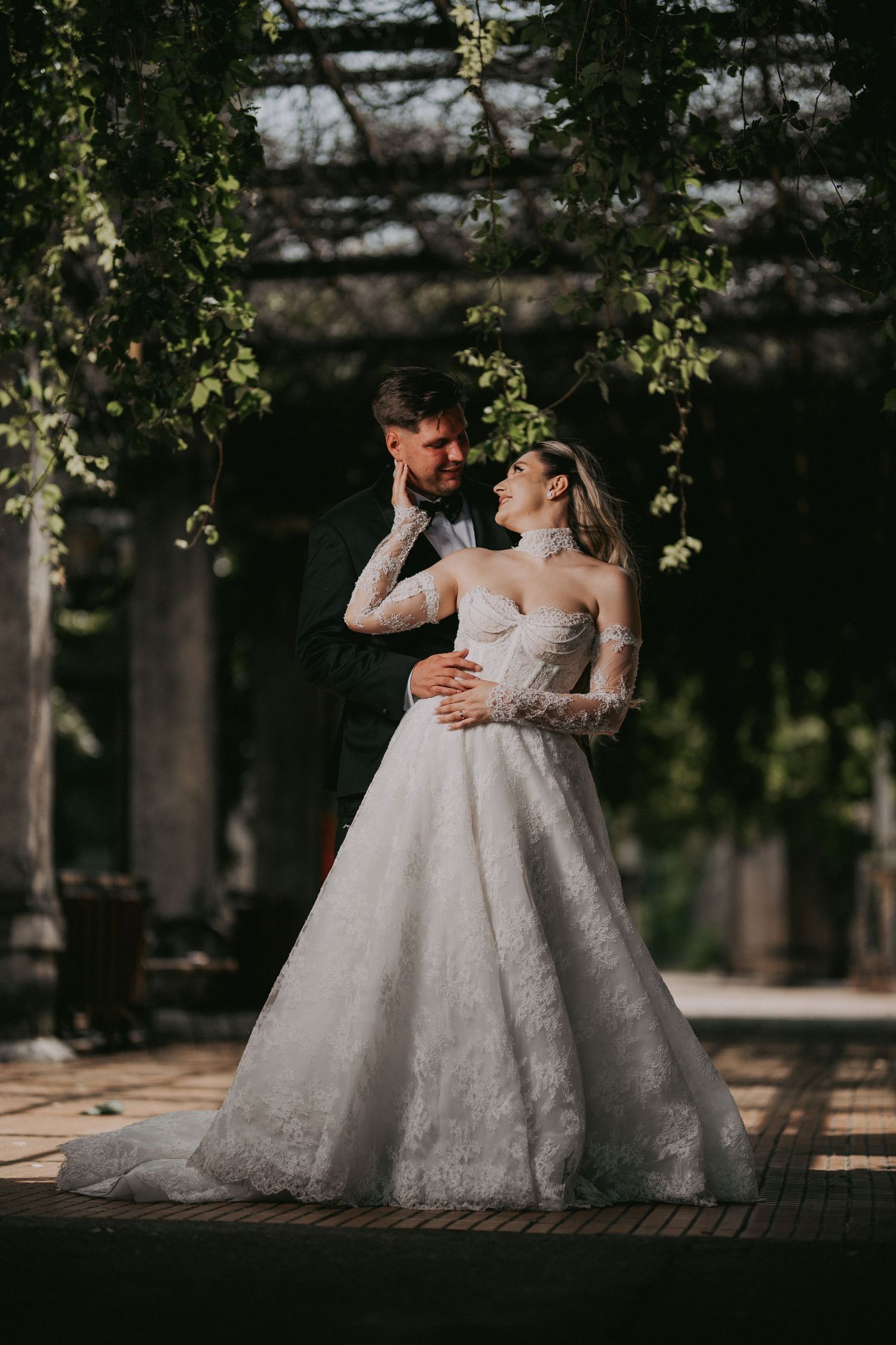 Bride and groom share an intimate embrace under a lush green pergola in soft sunlight.