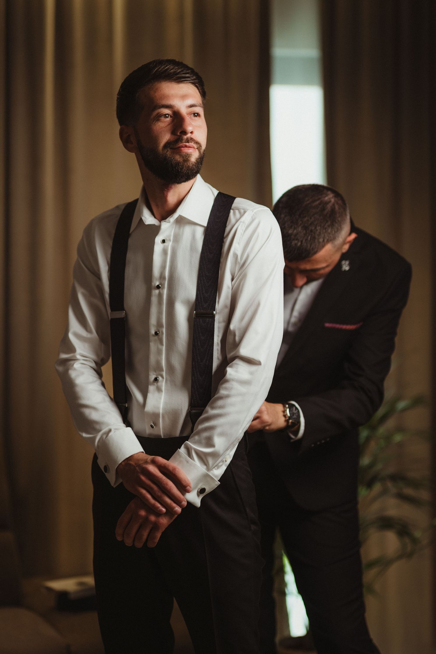 Groom in white shirt with suspenders being helped to dress by best man in a hotel room.