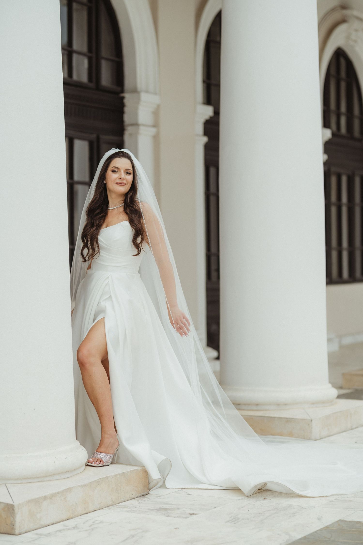 Beautiful bride in a strapless white wedding dress with veil and thigh slit posing between white columns.