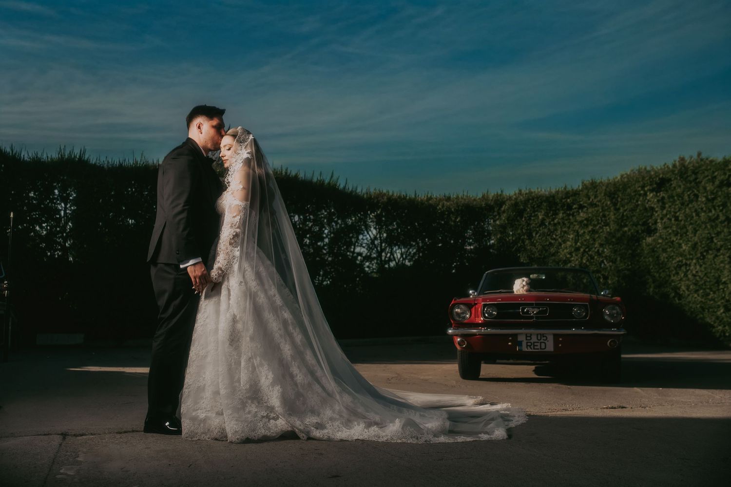 Bride and groom sharing romantic moment beside vintage red Mustang.