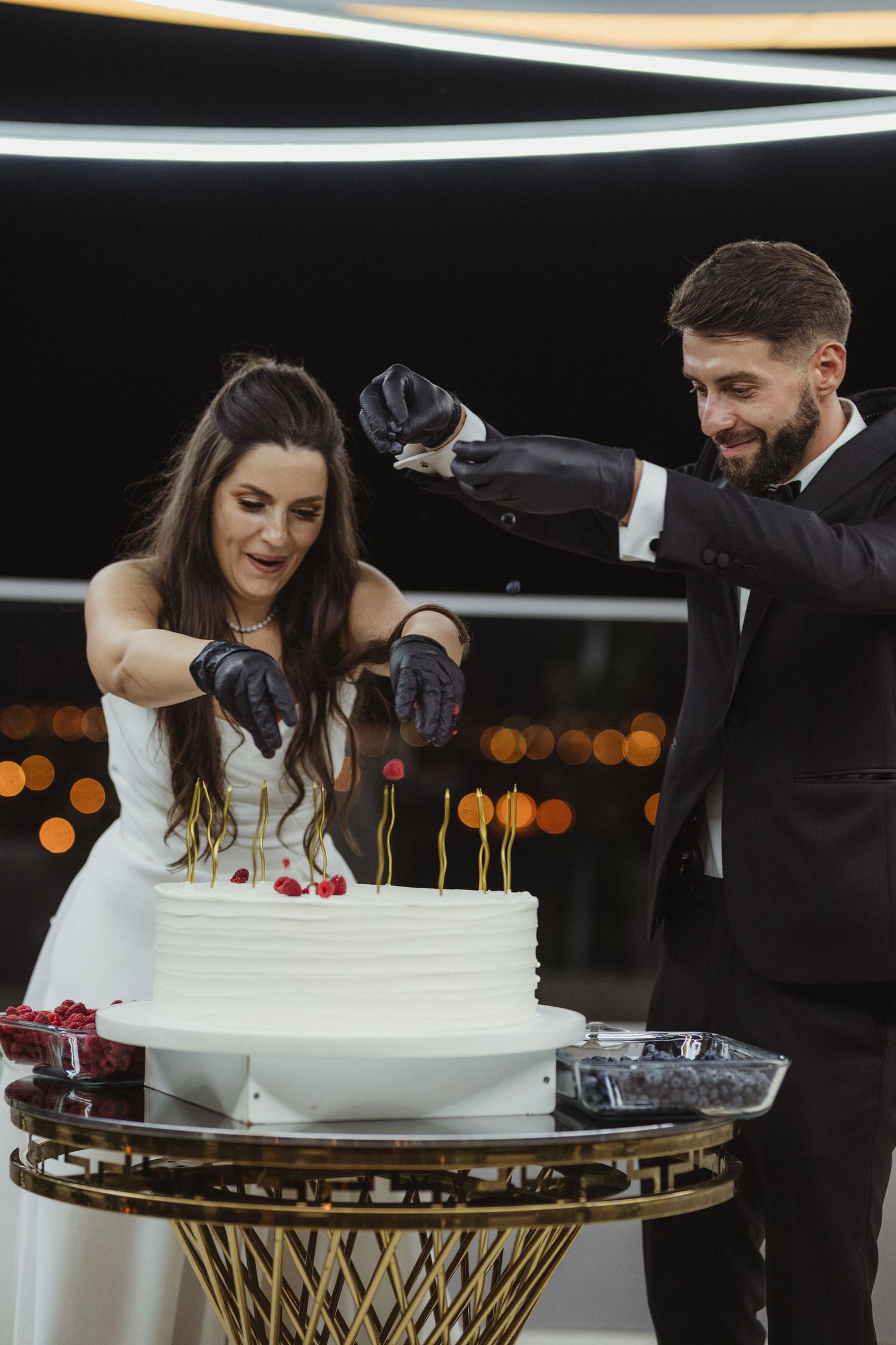 Bride and groom wearing black gloves cut elegant white wedding cake at nighttime reception.