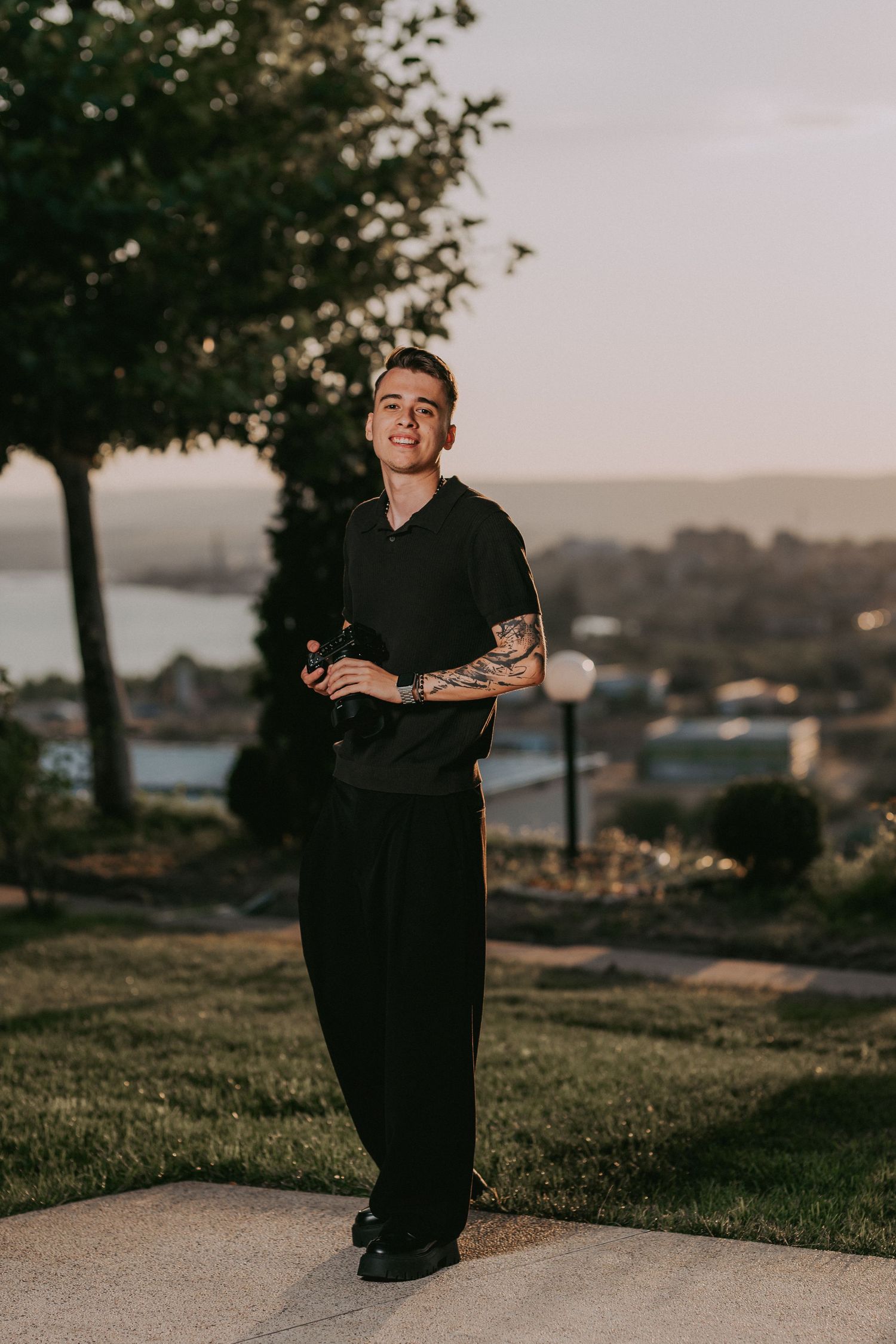 Tattooed young man in black outfit smiling confidently outdoors at sunset with scenic town view behind him. Epicu Fotograf Drobeta-Turnu Sev