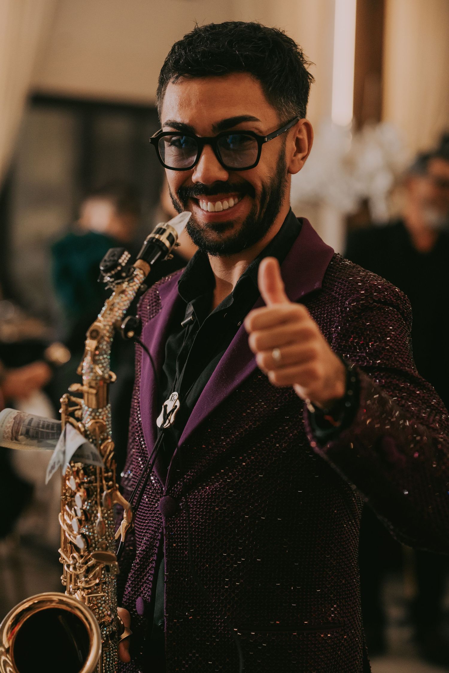 Smiling saxophonist in purple sequin jacket gives thumbs up while performing at a wedding reception.