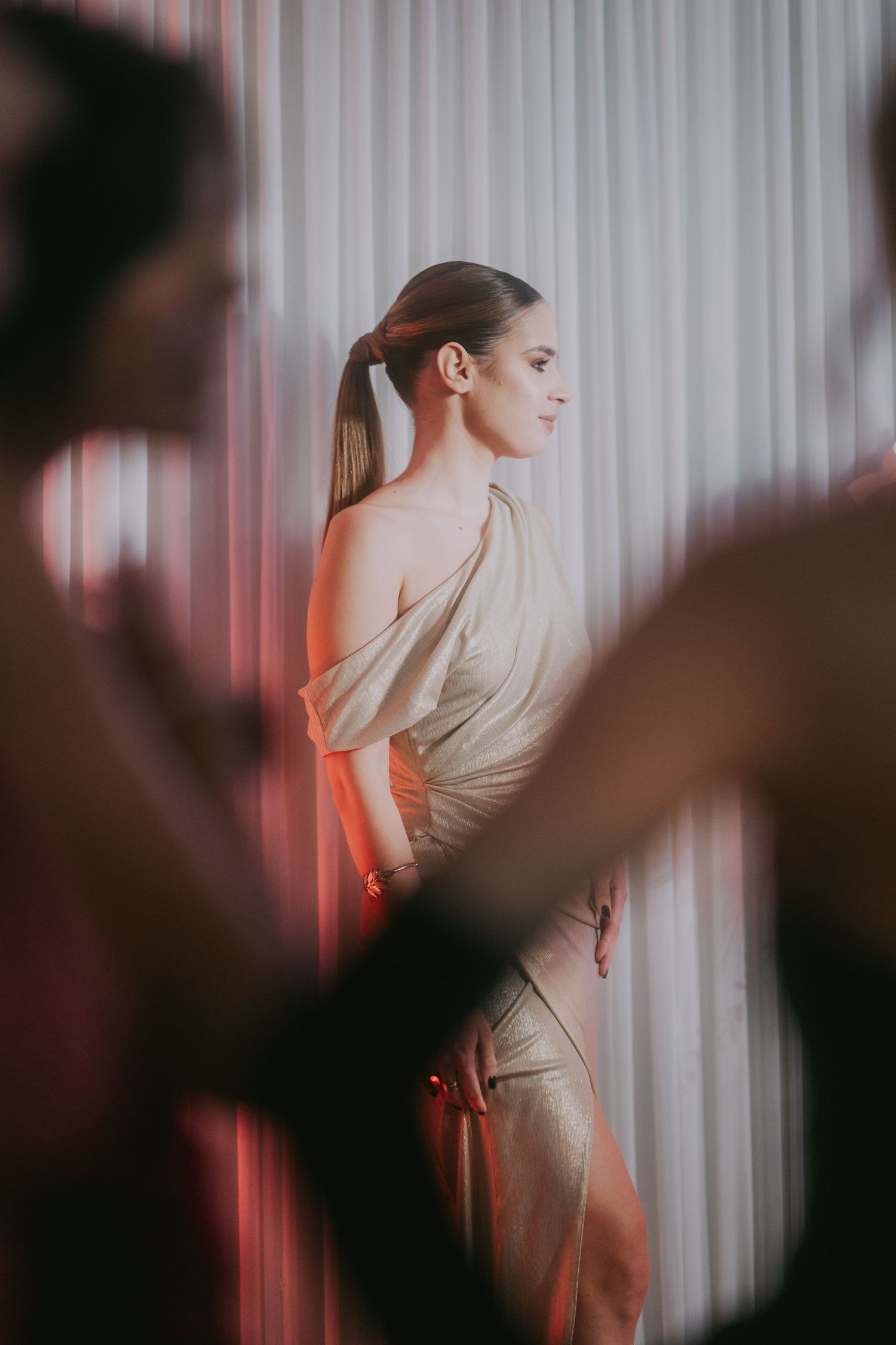 Elegant bride in gold one-shoulder gown with sleek ponytail standing by white curtains. Epicu Fotograf Drobeta-Turnu Severin