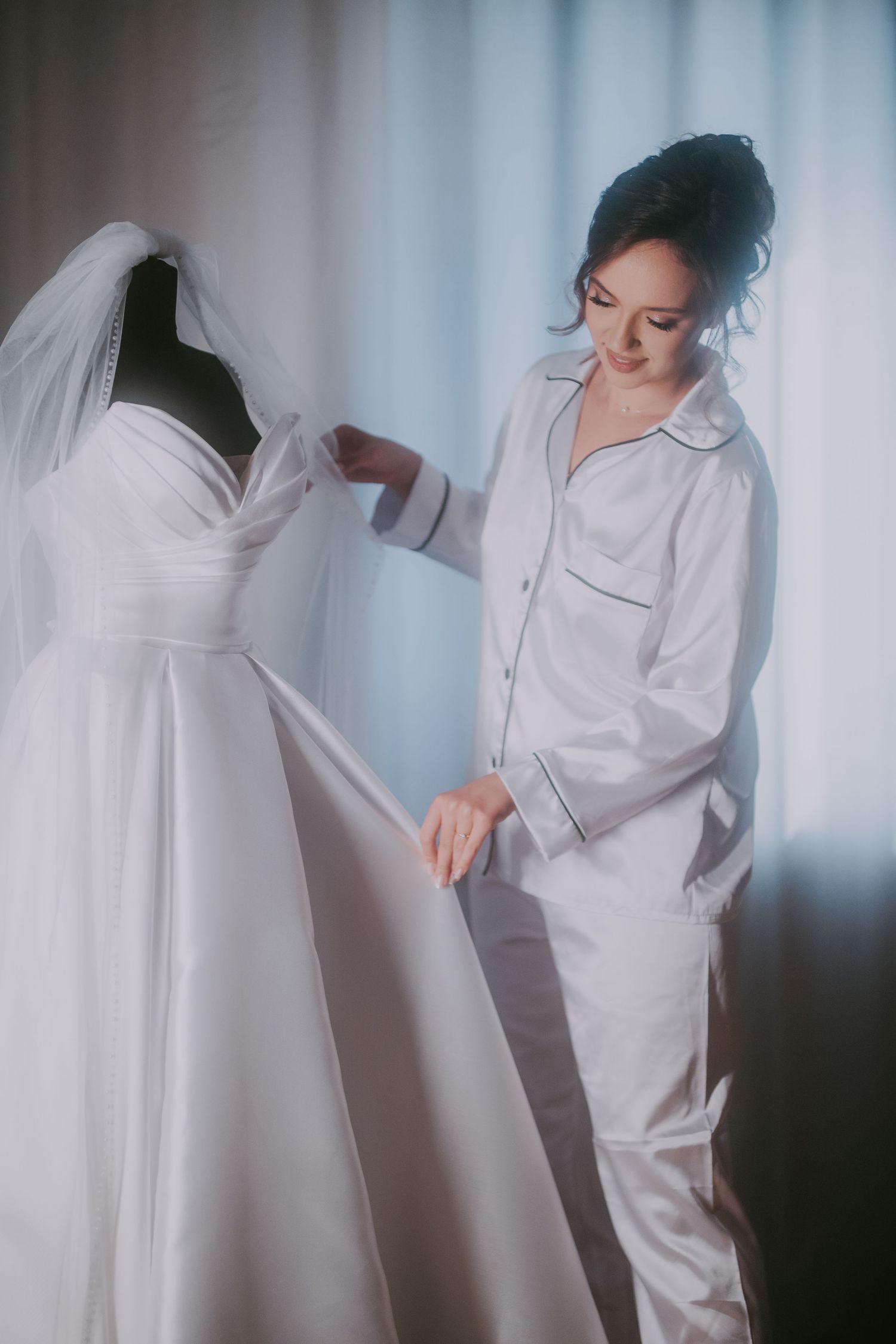 Bride in white satin pajamas admiring her wedding dress on a mannequin by a bright window. Epicu Fotograf Drobeta-Turnu Severin