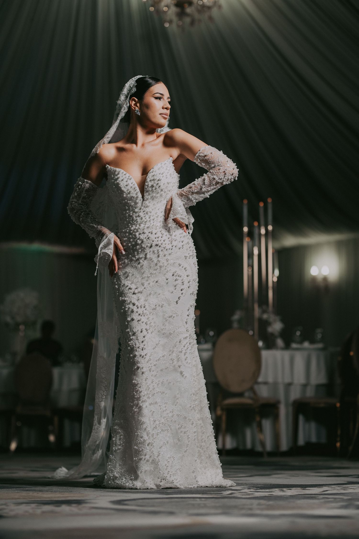 Bride in 3D floral beaded mermaid gown with veil poses under dramatic reception lighting.