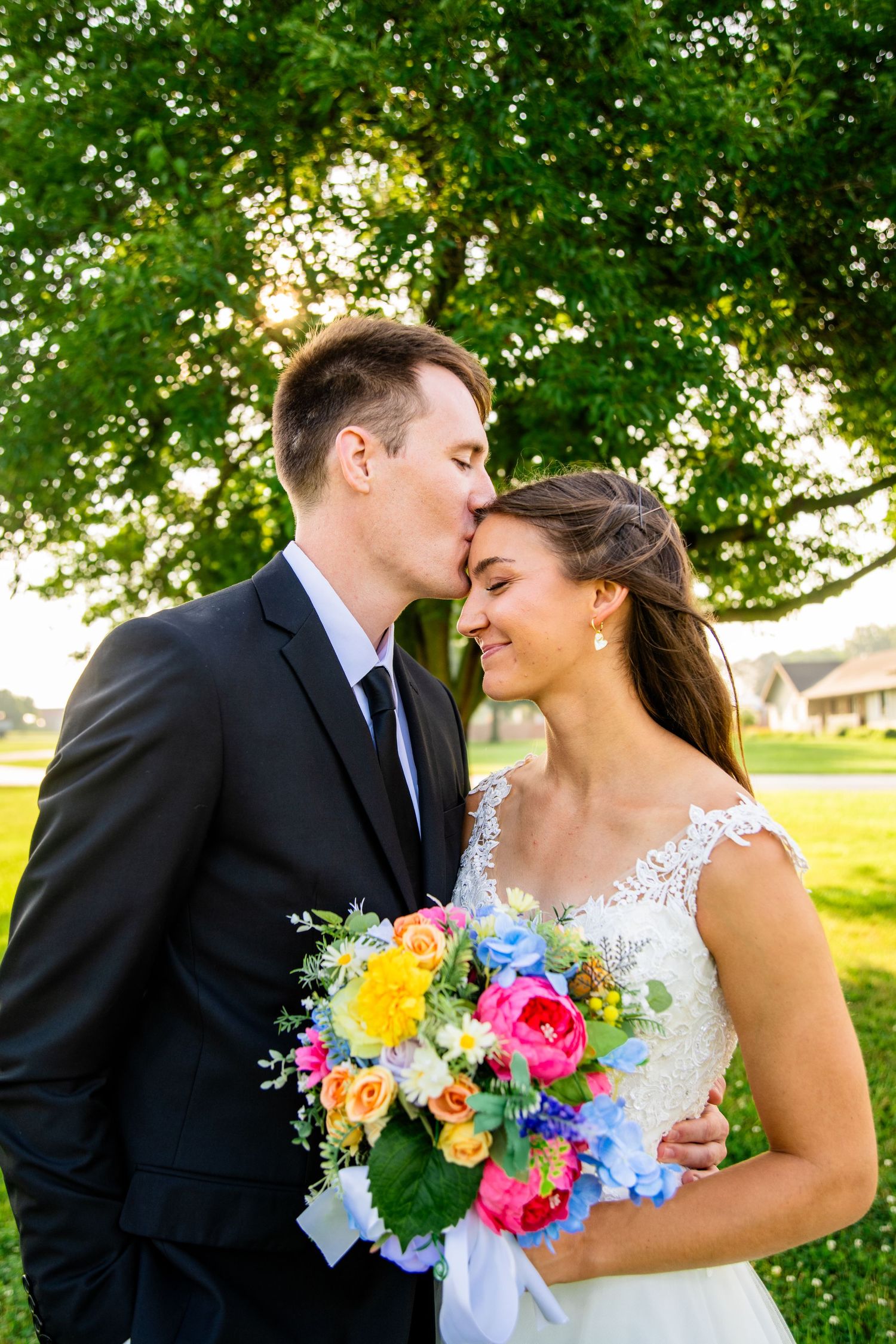 Groom kissing smiling bride on cheek while she holds a colorful wedding bouquet.