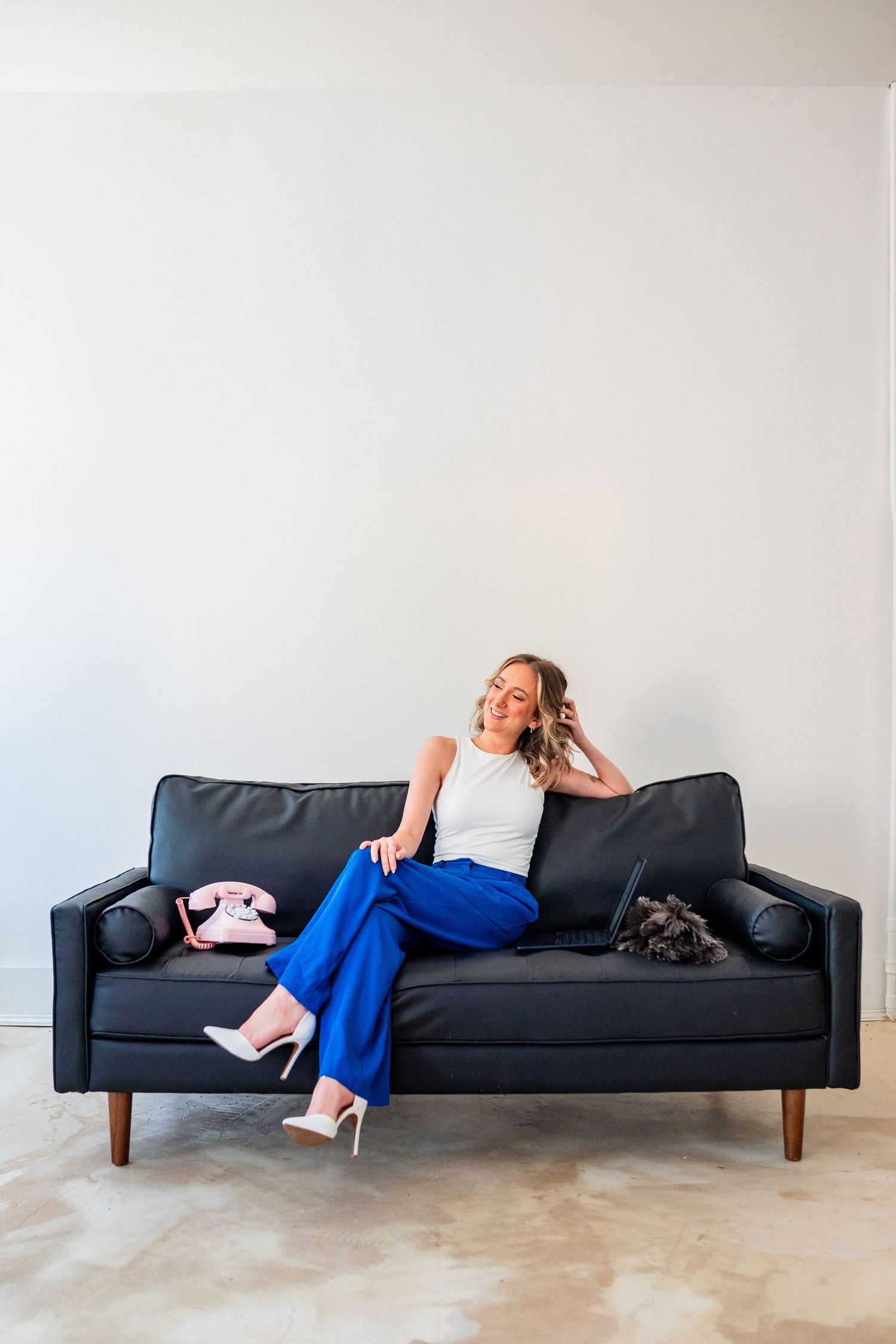 Woman in blue pants and white top relaxing on dark sofa with pink retro phone, smiling in bright minimalist room.