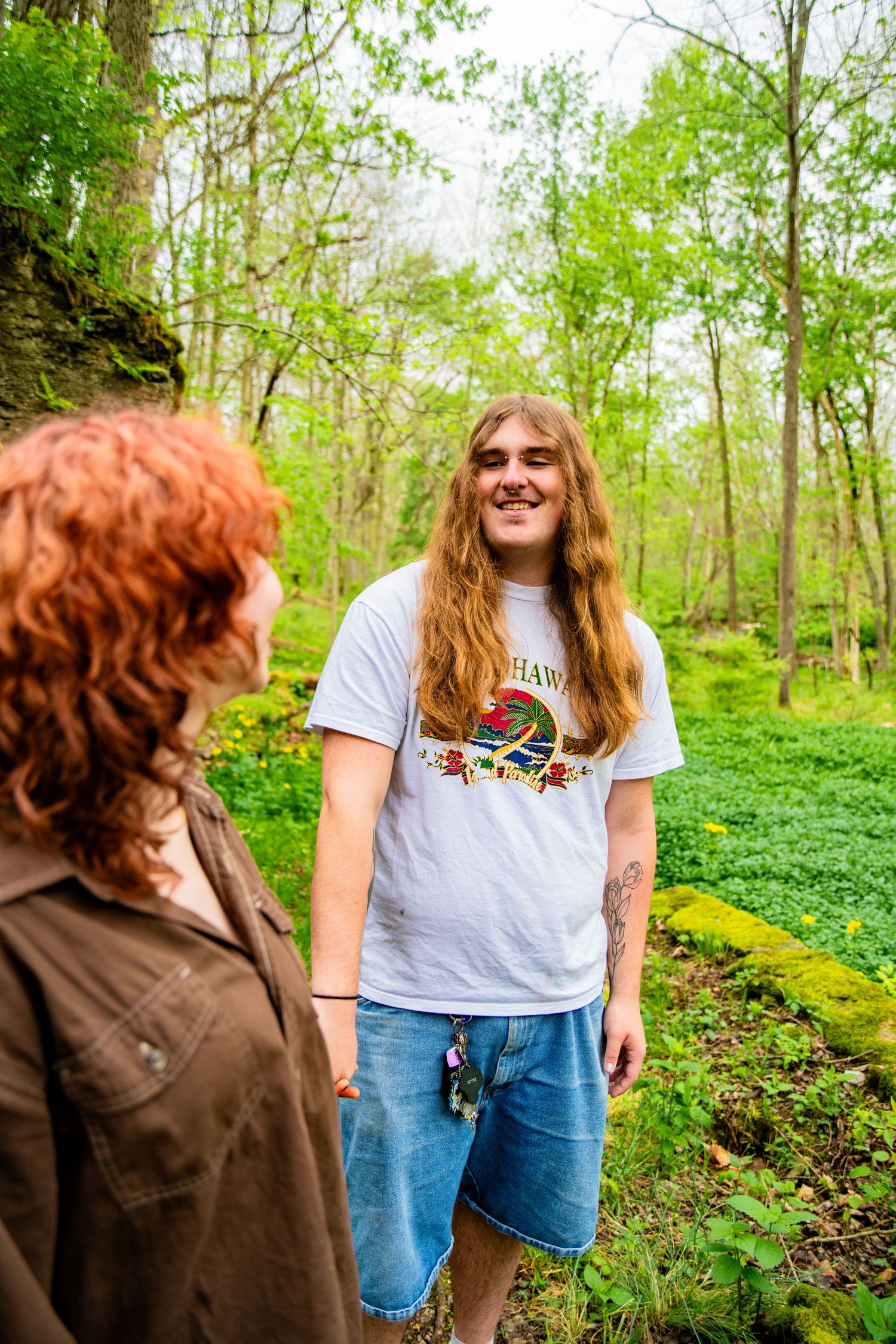 Two people with long hair conversing in a lush green forest, one wearing a white graphic tee and denim shorts.