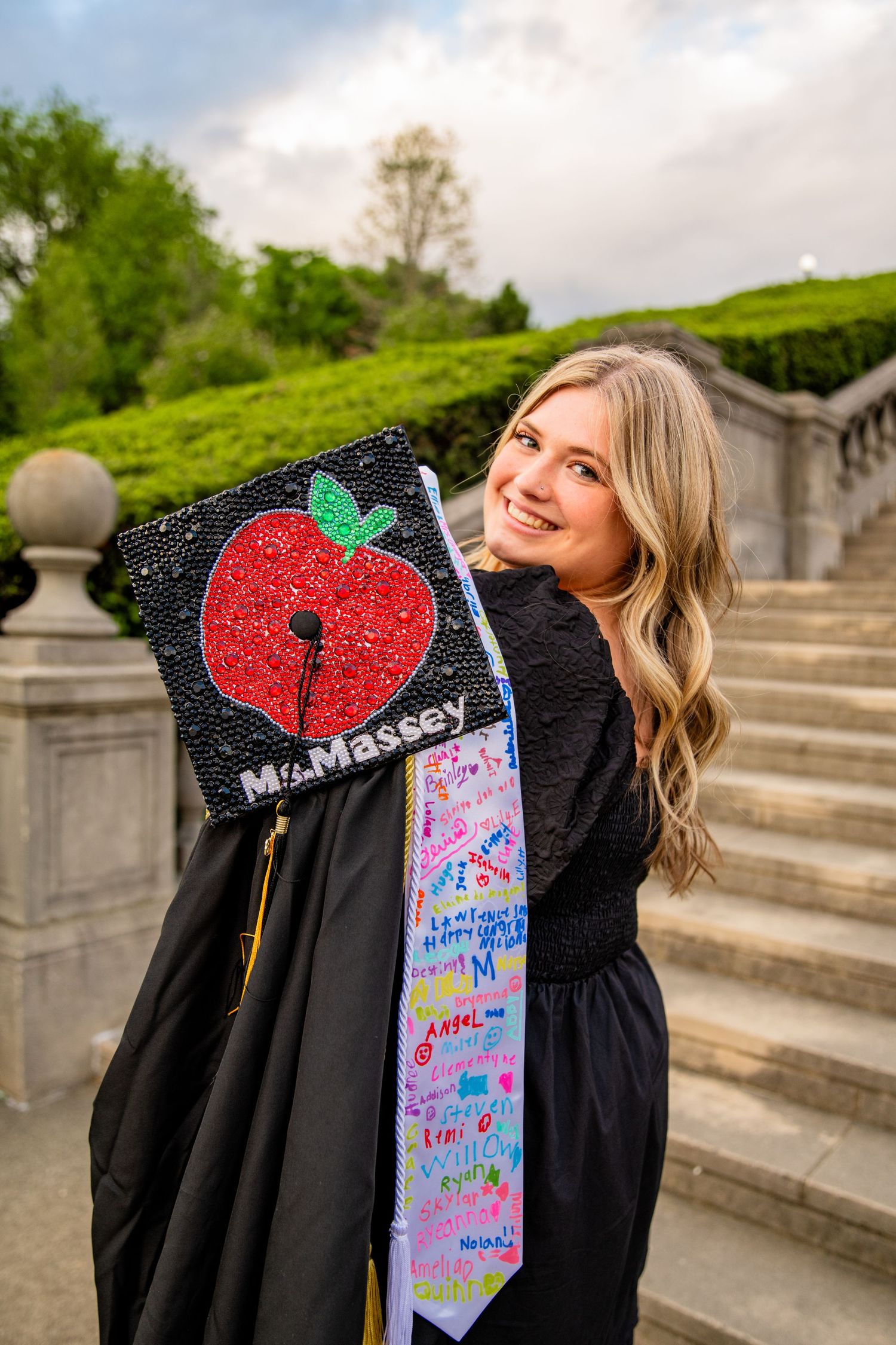 Smiling female graduate in black gown displays decorated cap reading Ms. Massey with red apple design on stone steps.