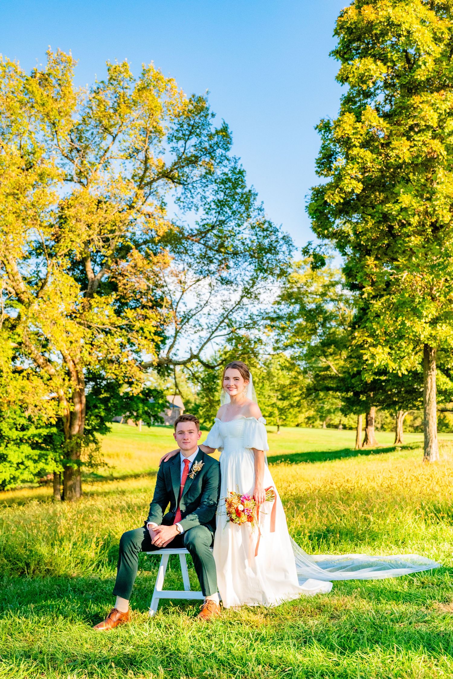 Bride and groom pose outdoors in autumn park, groom seated on white stool, bride holding floral bouquet.